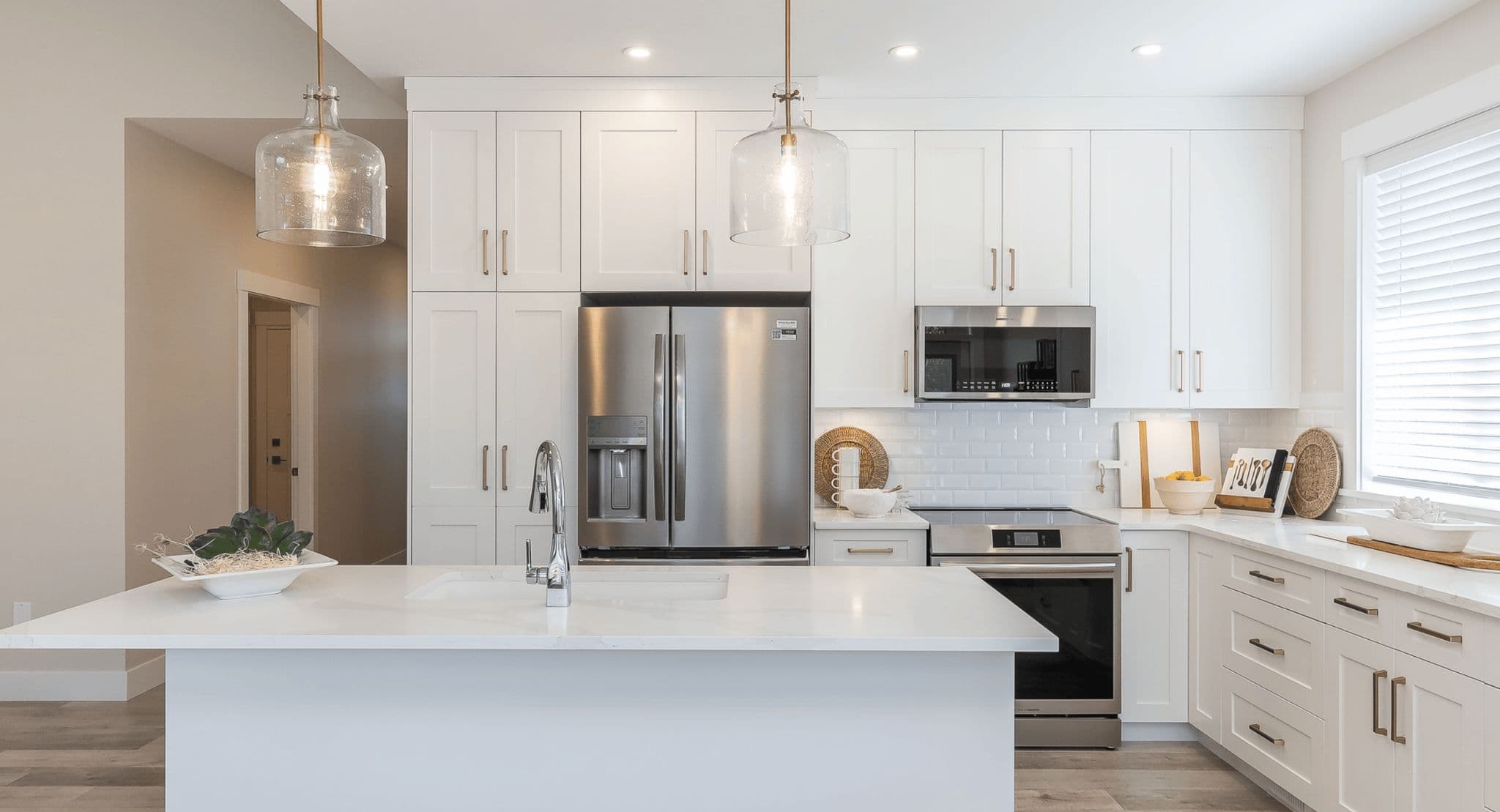 Malloway Village – A bright, modern kitchen featuring white shaker-style cabinetry, white quartz countertops, and stainless steel appliances including a French-door refrigerator, range, and over-the-range microwave. A central kitchen island with an undermount sink and chrome faucet is illuminated by two glass pendant lights, complemented by a white subway tile backsplash.
