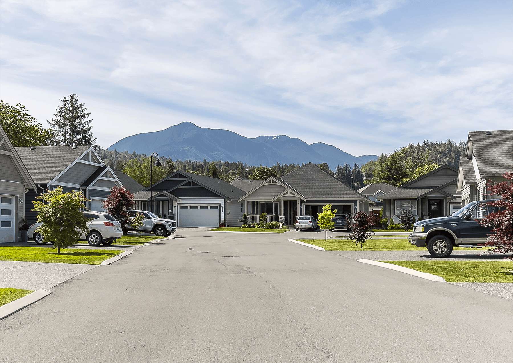 Malloway Village – A paved suburban cul-de-sac lined with modern craftsman-style single-family homes, manicured green lawns, and parked cars under a bright daytime sky. Large, forested mountains rise prominently directly behind the neighborhood in the background.