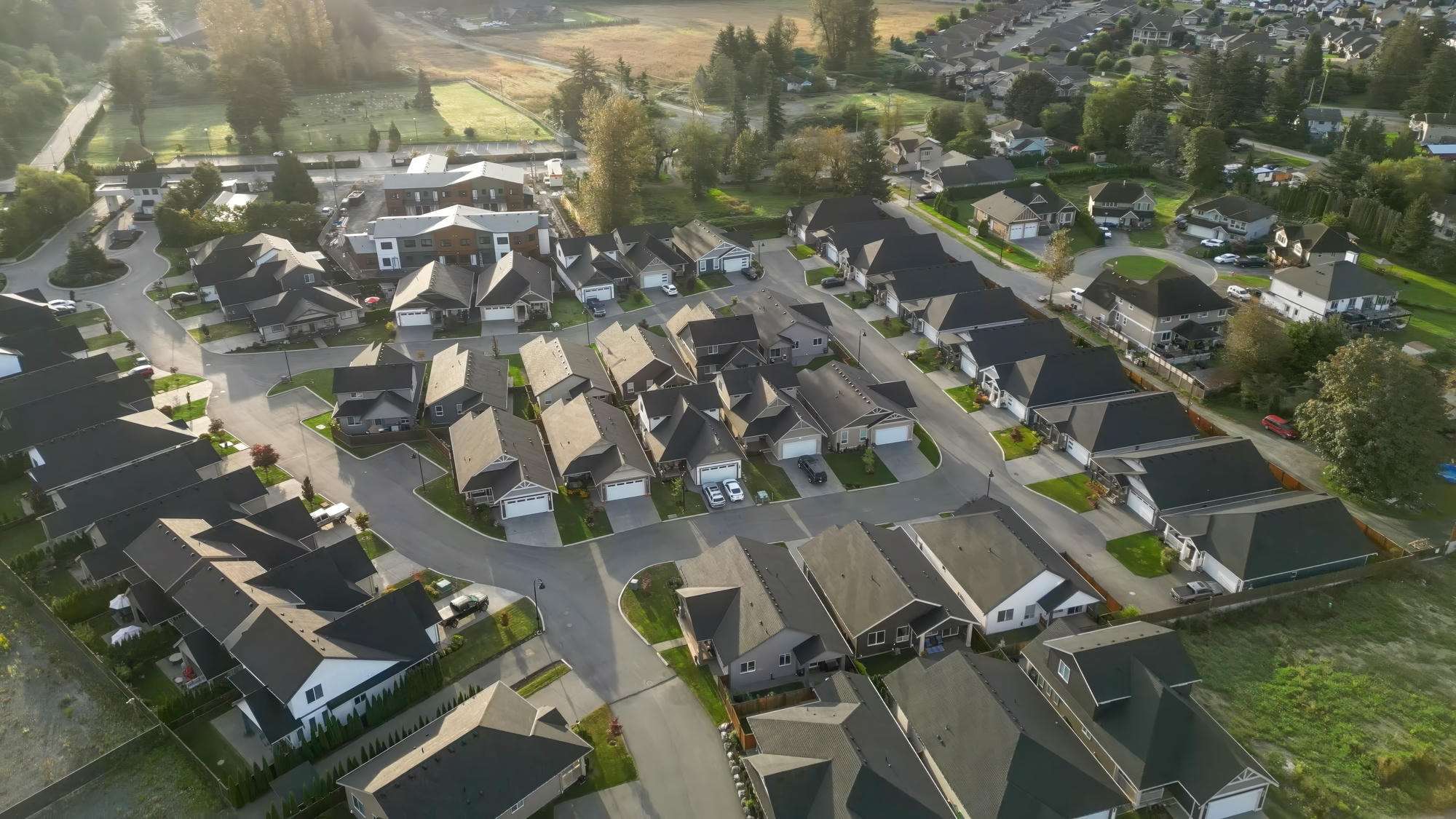 Malloway Village – An elevated drone view captures a planned suburban subdivision of uniform, single-family craftsman-style homes with dark shingled roofs and paved cul-de-sacs. The surrounding neighborhood borders green fields and scattered mature trees bathed in soft, early-morning sunlight.