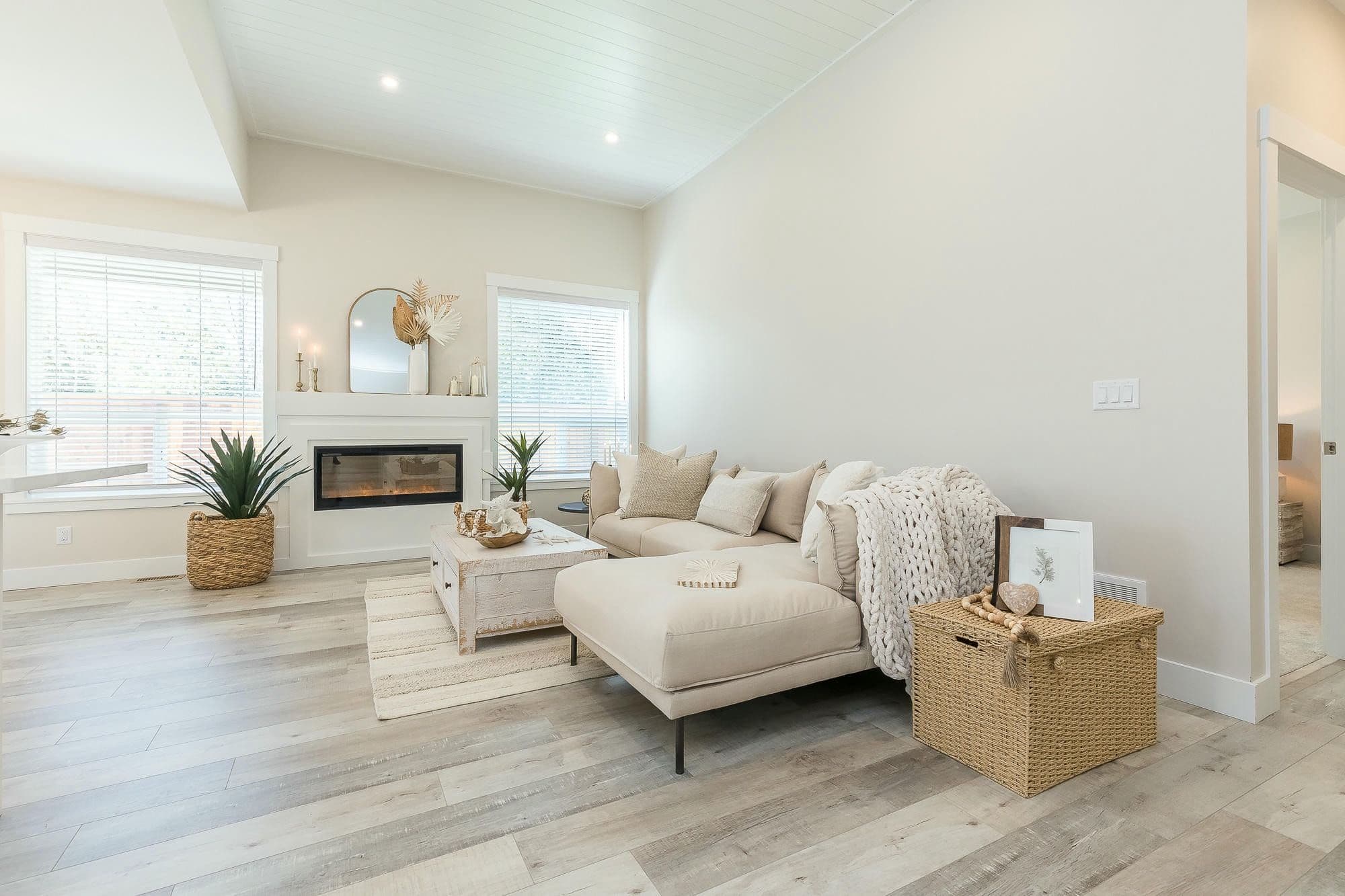 Malloway Village – A bright, neutrally styled living room features light wood-look flooring, high angled ceilings with recessed lighting, and a modern built-in fireplace flanked by two windows with blinds. The space is furnished with an off-white sectional sofa with a chaise, a whitewashed coffee table, woven storage baskets, and textured boho-chic decor accents.