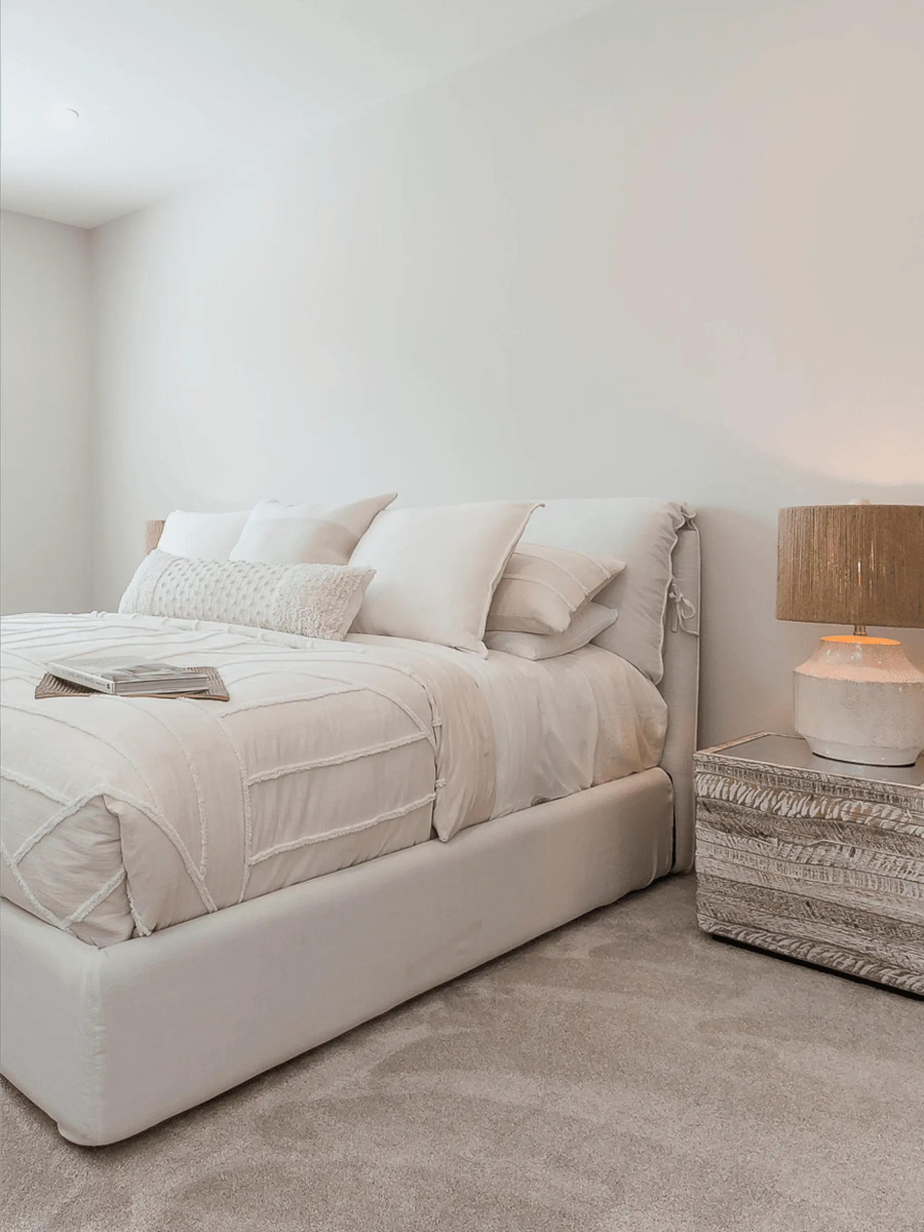 Malloway Village – A bright, neutral bedroom featuring an upholstered bed dressed in textured cream linens and an abundance of matching pillows. Beside the bed sits a weathered whitewashed wooden nightstand topped with an earthy ceramic and rattan lamp casting warm light against the off-white wall.