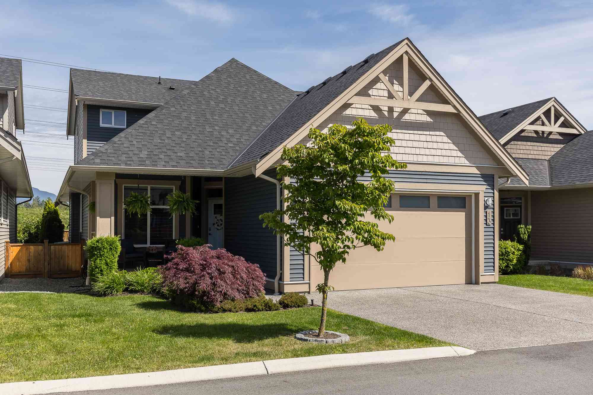 Malloway Village – A modern craftsman-style townhouse featuring blue-grey horizontal siding, shake-shingle and timber gable accents, and an attached beige garage. The home is fronted by a paved driveway, a manicured green lawn, a young deciduous tree, and low decorative shrubbery under a clear blue sky.