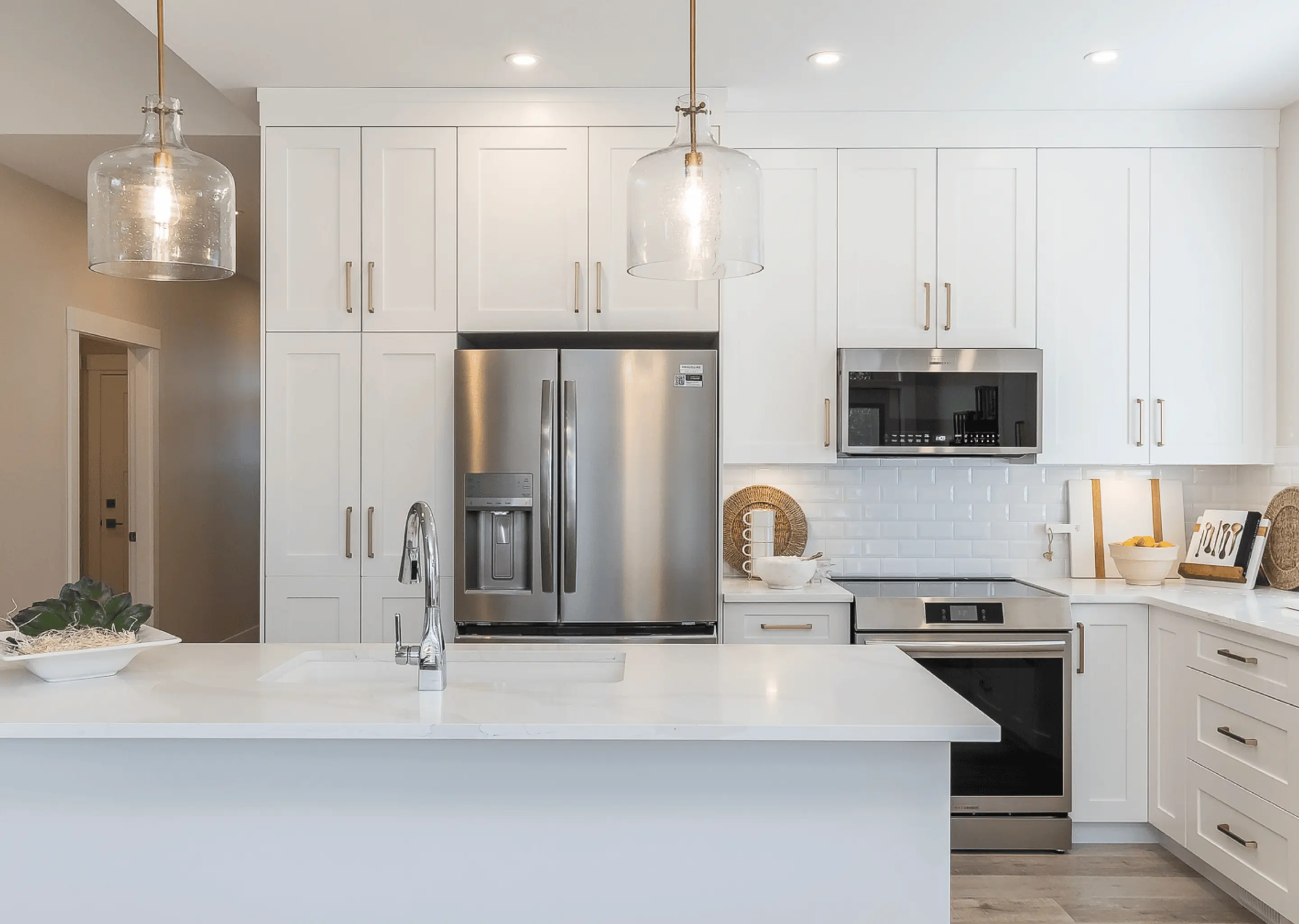 Malloway Village – A bright modern kitchen features white shaker cabinets, a matching white island with an undermount sink and chrome faucet, and stainless steel appliances including a French-door refrigerator, range, and microwave. Large glass cloche pendant lights hang above the island against a white subway tile backsplash and brass cabinet hardware.