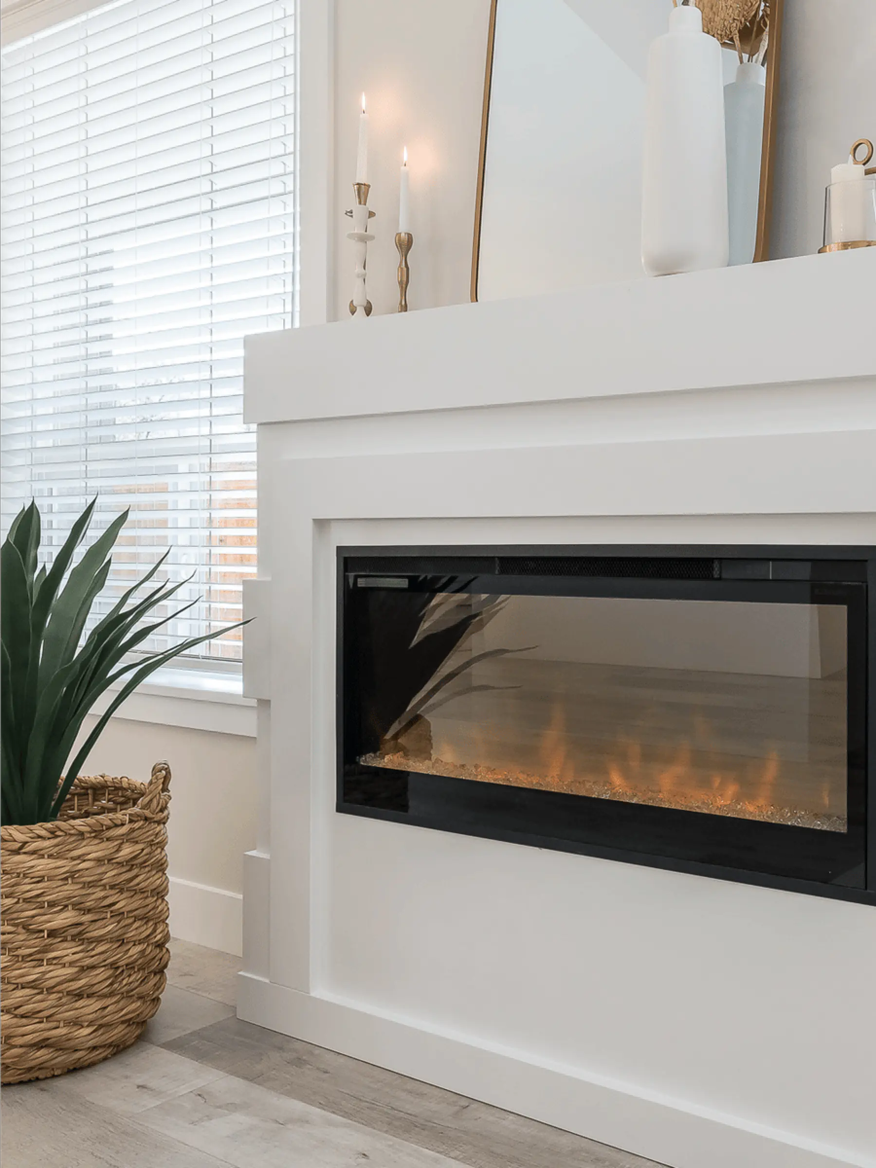Malloway Village – A modern living space featuring a built-in electric fireplace set inside a white-painted mantel structure. A potted snake plant in a woven basket sits to the left next to a window with horizontal blinds, while lit candles and decorative vases adorn the mantelpiece.