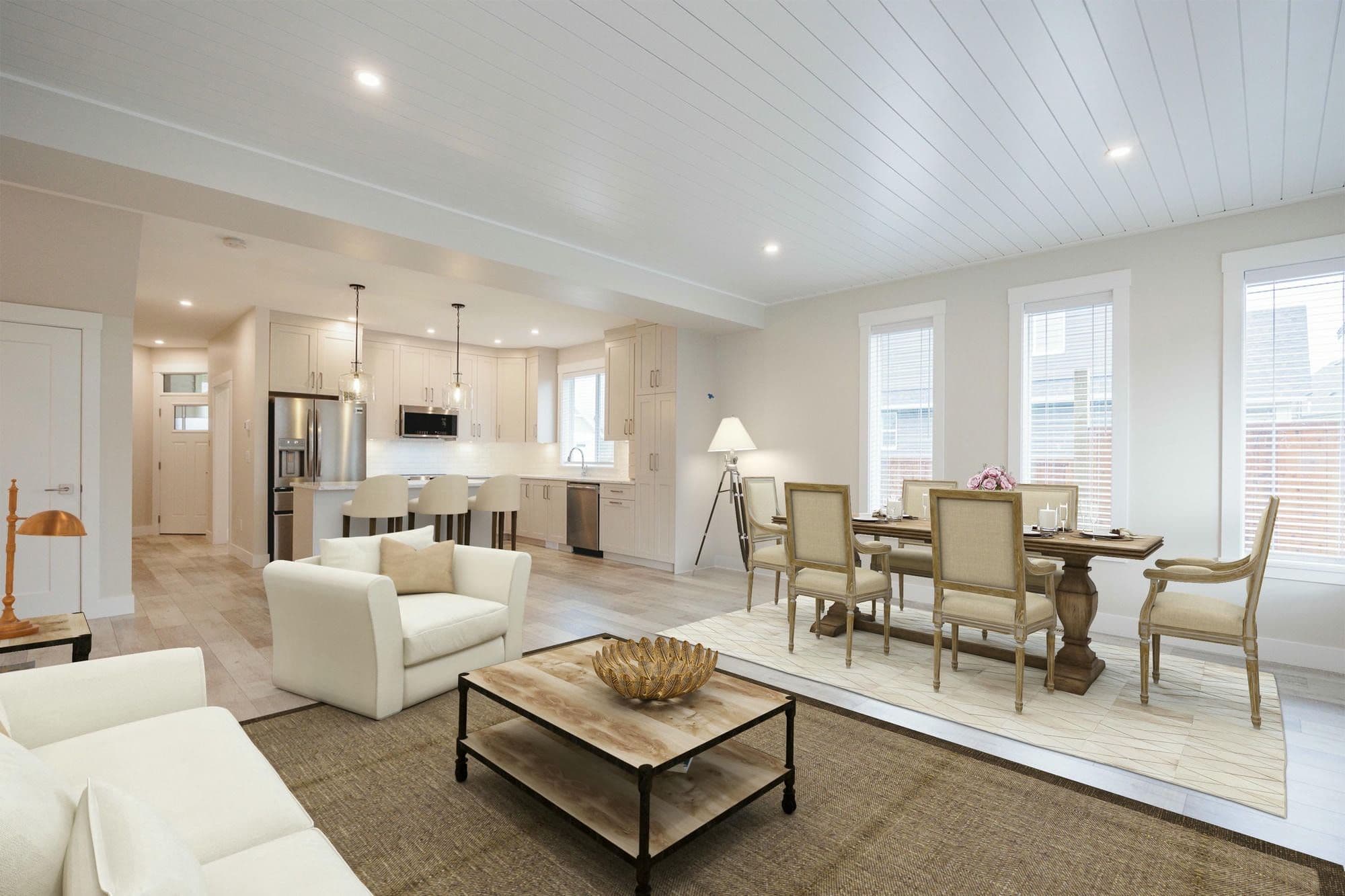 Malloway Village – An open-concept living, dining, and kitchen area featuring white-panelled ceilings, light-coloured walls, and light wood flooring. A living setup with cream seating anchors the foreground next to a traditional dining table set with upholstered chairs, while the background reveals a modern kitchen with white cabinetry and stainless steel appliances.