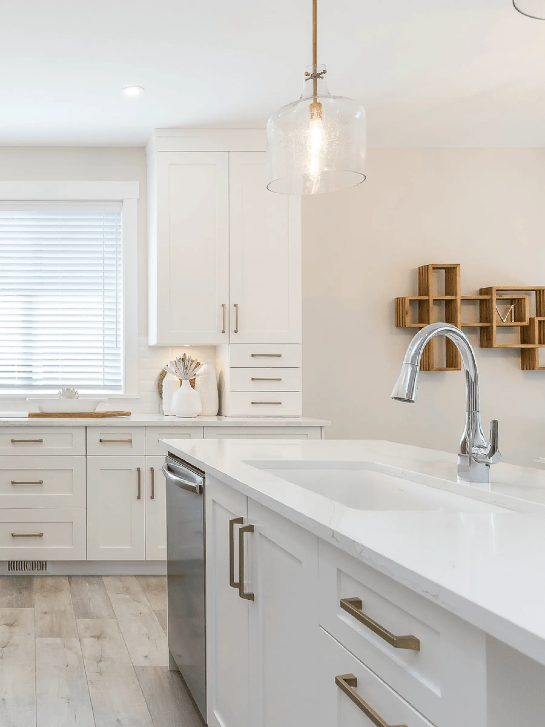 Malloway Village – A bright, modern kitchen featuring a white quartz-topped island with an undermount sink and chrome gooseneck faucet. Shaker-style white cabinetry with brass handles, light wood flooring, and a glass bell pendant light create a clean, contemporary ambiance.