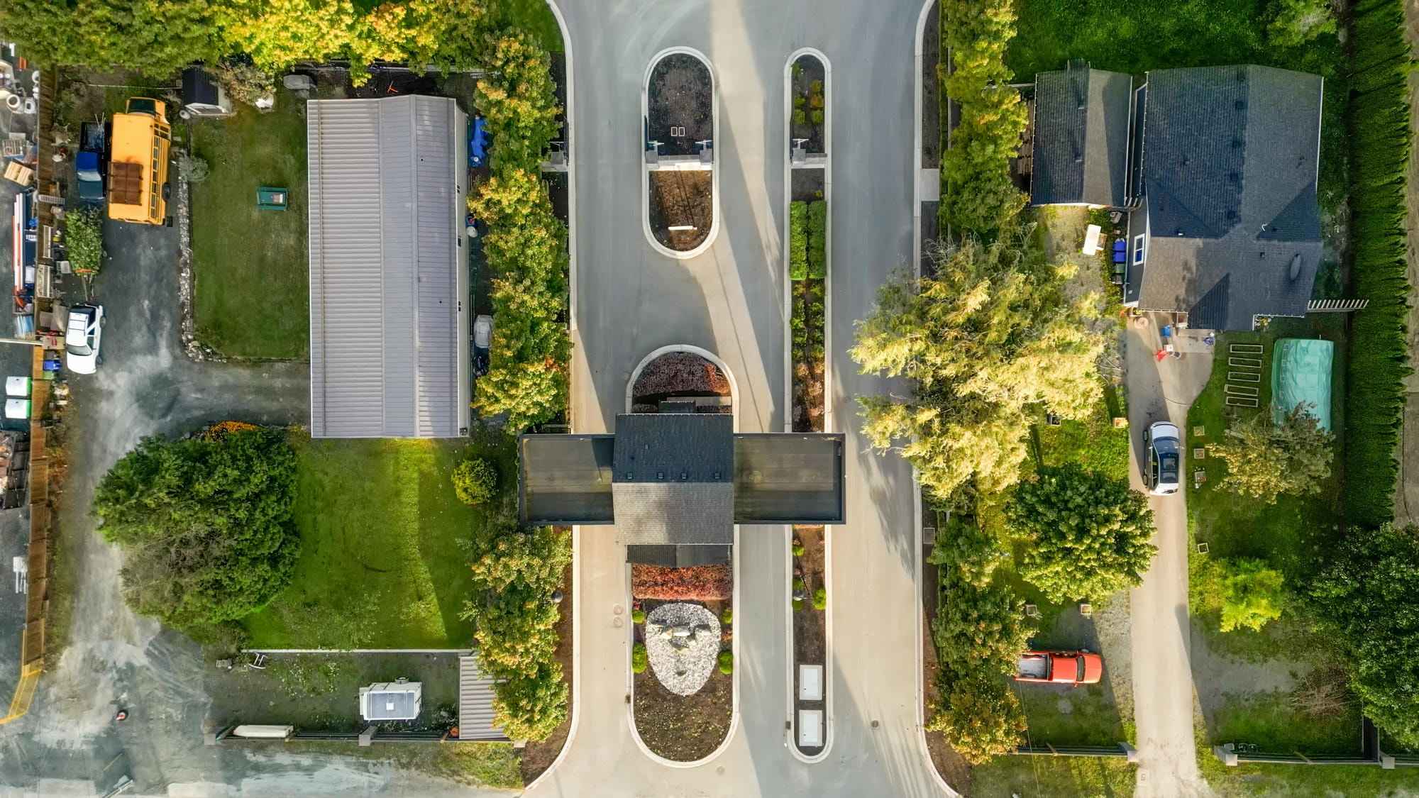 Malloway Village – A top-down aerial view of a gated community entrance with a central security gatehouse and paved multi-lane roadway. The entrance is framed by landscaped medians, lush green lawns, mature trees, and neighboring residential homes under bright daytime light.