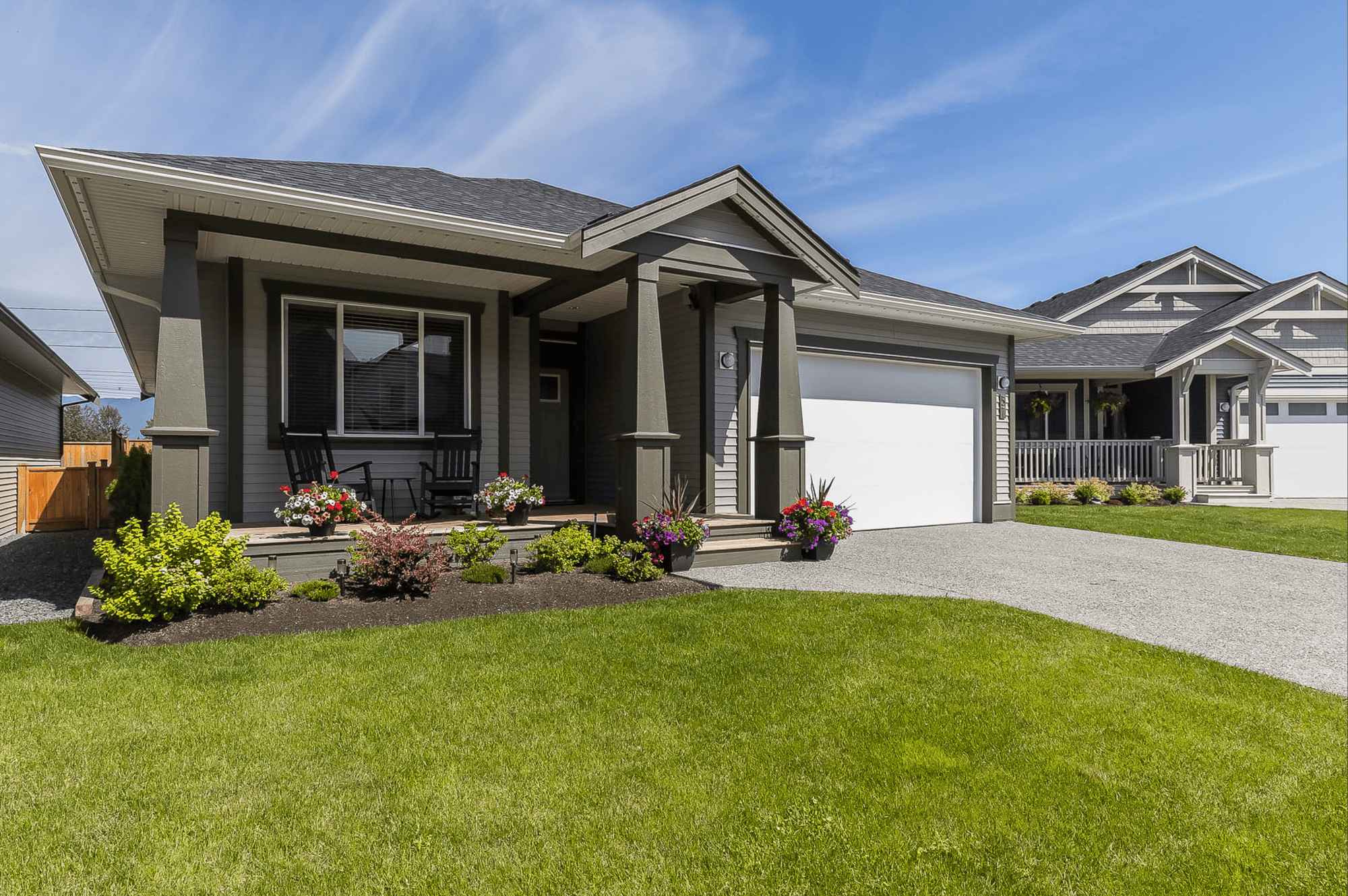Malloway Village – A ground-level view of a single-story craftsman-style house featuring grey siding, dark green trim and columns, and a white garage door. A neat green lawn, landscaped flower beds, and a concrete driveway sit in the foreground under a bright blue sky with light wispy clouds.