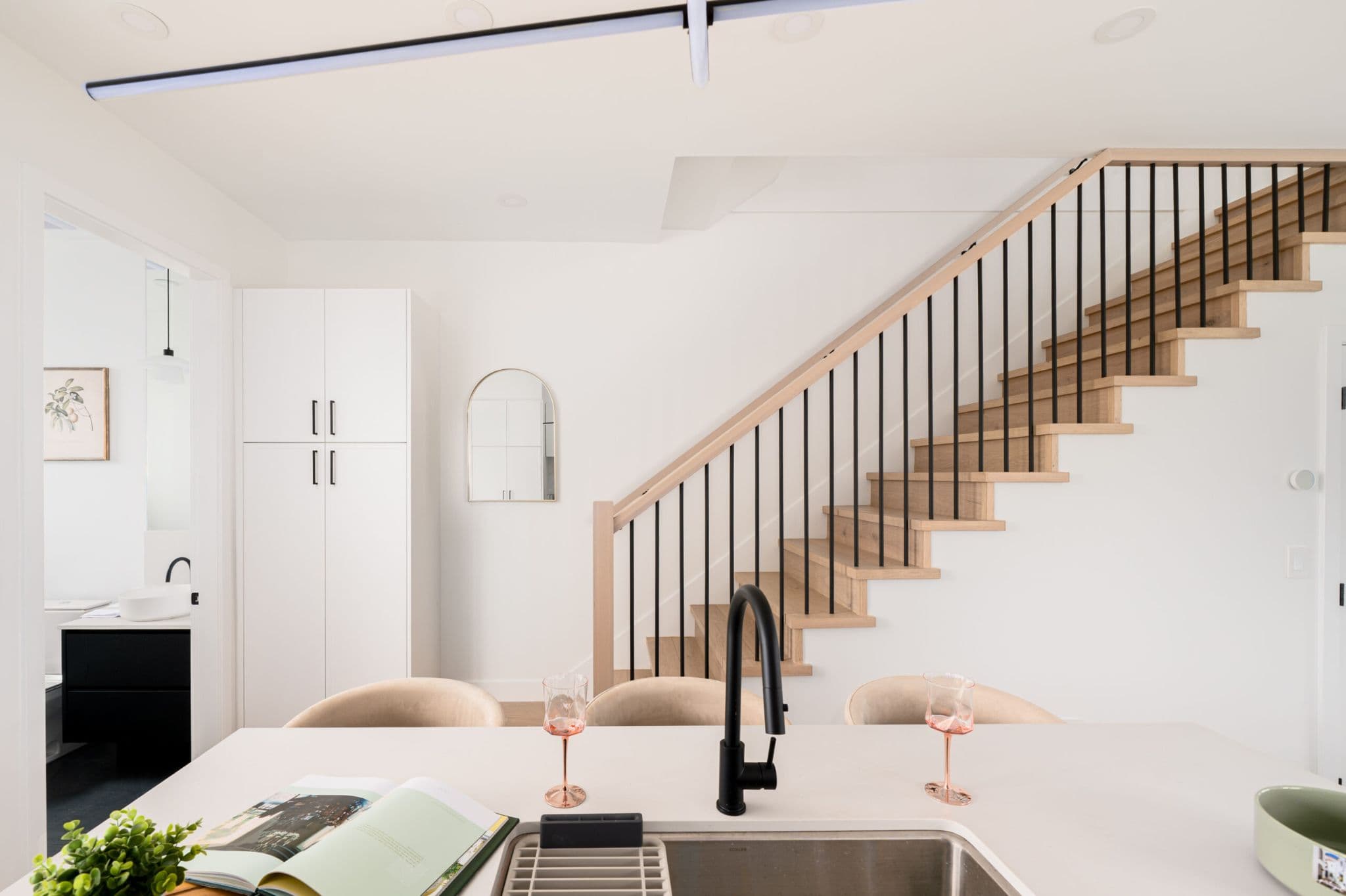 August Laurel + W64 – A modern interior view looking across a kitchen island toward a light-wood staircase with black metal spindles and crisp white walls. In the foreground, the counter features a matte black faucet, wine glasses, and an open magazine, while a bathroom and white cabinetry are visible in the background.