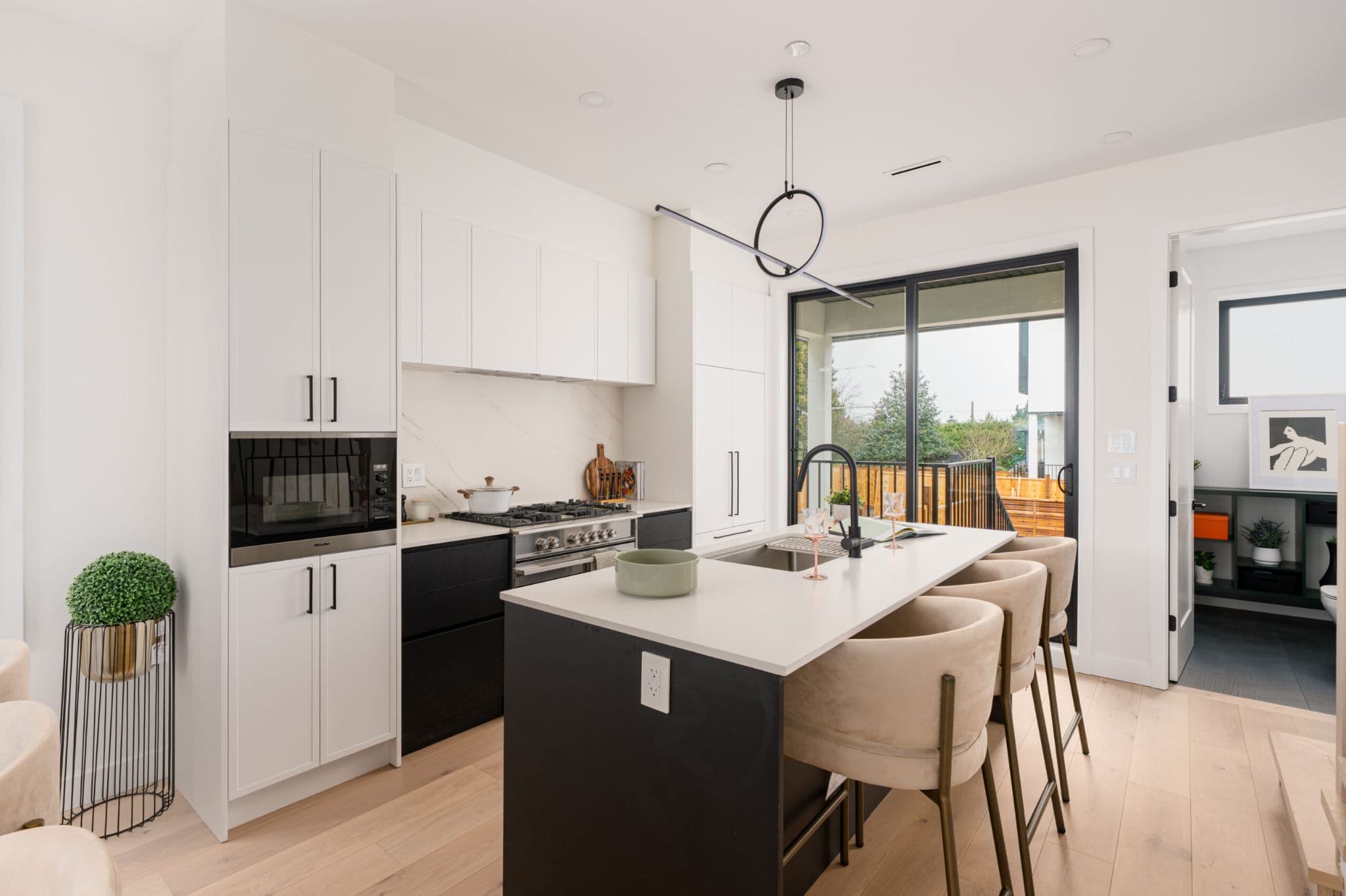 August Laurel + W64 – A modern open-concept kitchen featuring a dark island with white quartz countertops, paired with three upholstered beige barstools and a minimalist ring pendant light above. The space includes white upper cabinetry, dark lower drawers, integrated appliances, light hardwood floors, and sliding glass doors that open to an outdoor patio.