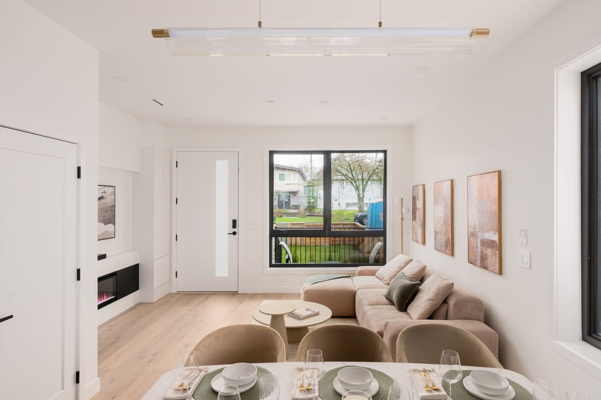 August Laurel + W64 – A modern open-concept living room and dining space features light hardwood floors, crisp white walls, and an L-shaped beige sectional sofa. A large black-framed window looks out onto a green front yard and residential street, while a contemporary linear light fixture hangs above the staged dining table in the foreground.