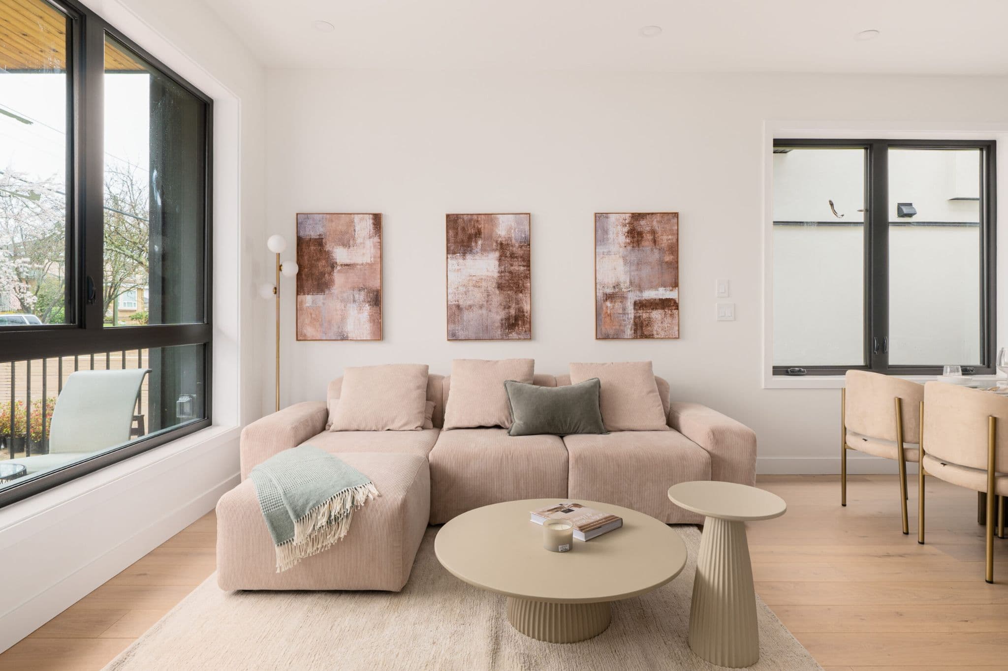 August Laurel + W64 – A modern living room features a plush beige sectional sofa adorned with three framed abstract artworks mounted on the white wall behind it. Large black-framed windows bring in ample natural light, highlighting light hardwood floors, a round coffee table, and an adjacent dining area.