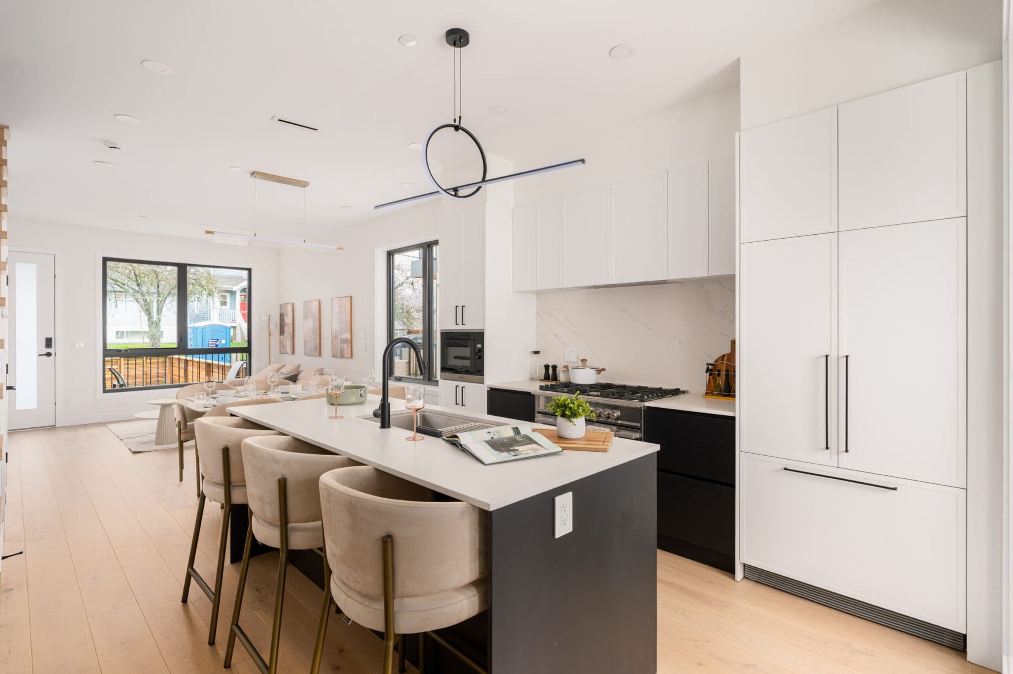 August Laurel + W64 – A modern open-concept kitchen and dining area featuring a dark kitchen island with a white countertop, beige upholstered bar stools, and integrated white cabinetry. A sleek circular black pendant light hangs above the island, leading the eye toward the adjoining dining space bathed in natural light from large windows.