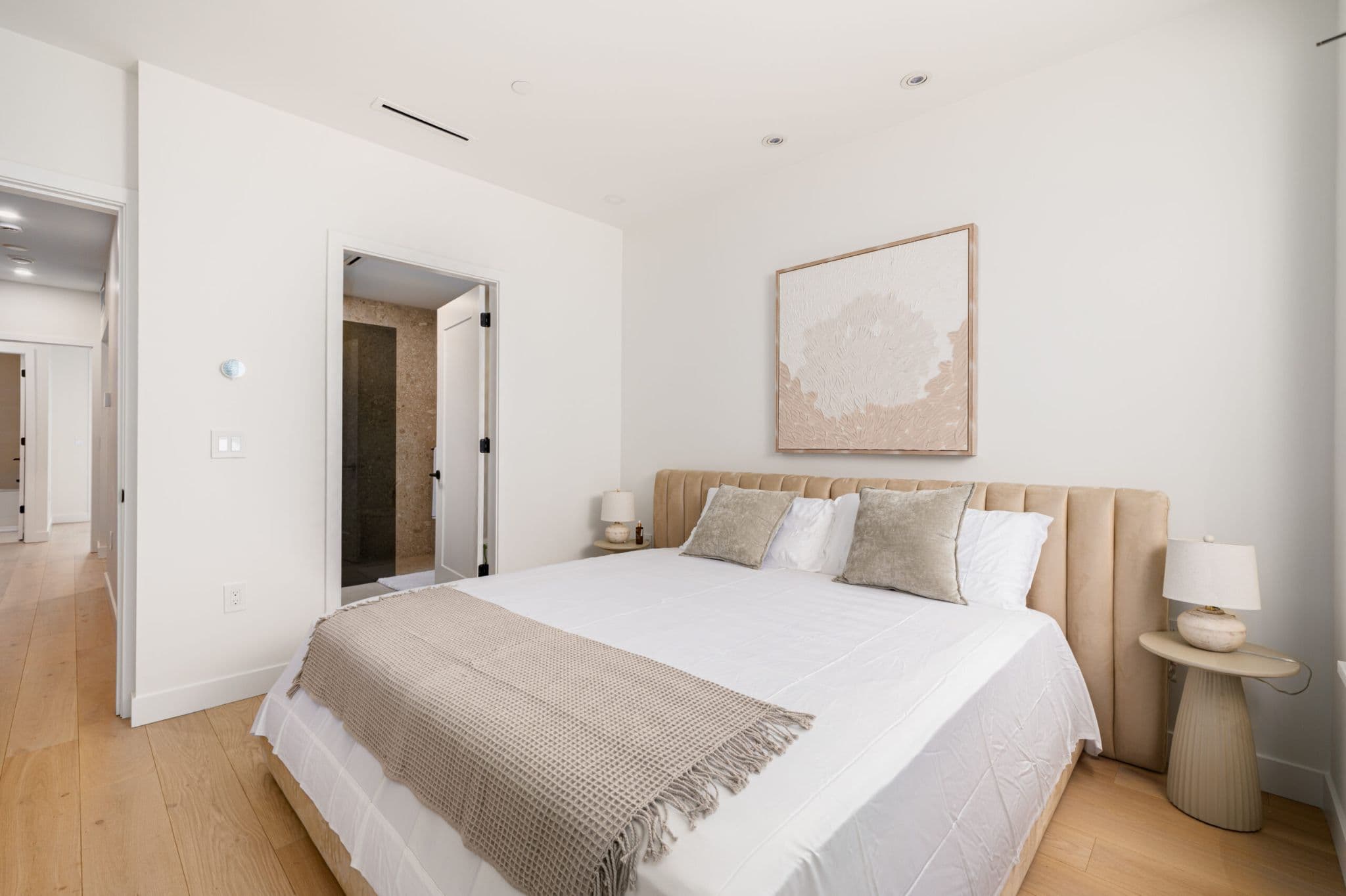 August Laurel + W64 – A modern, bright bedroom features a king-sized bed with a beige tufted headboard, crisp white linens, and neutral accent pillows. Textured artwork hangs on the white wall above the bed, with light wood flooring extending toward a doorway that reveals a stone-tiled ensuite bathroom.