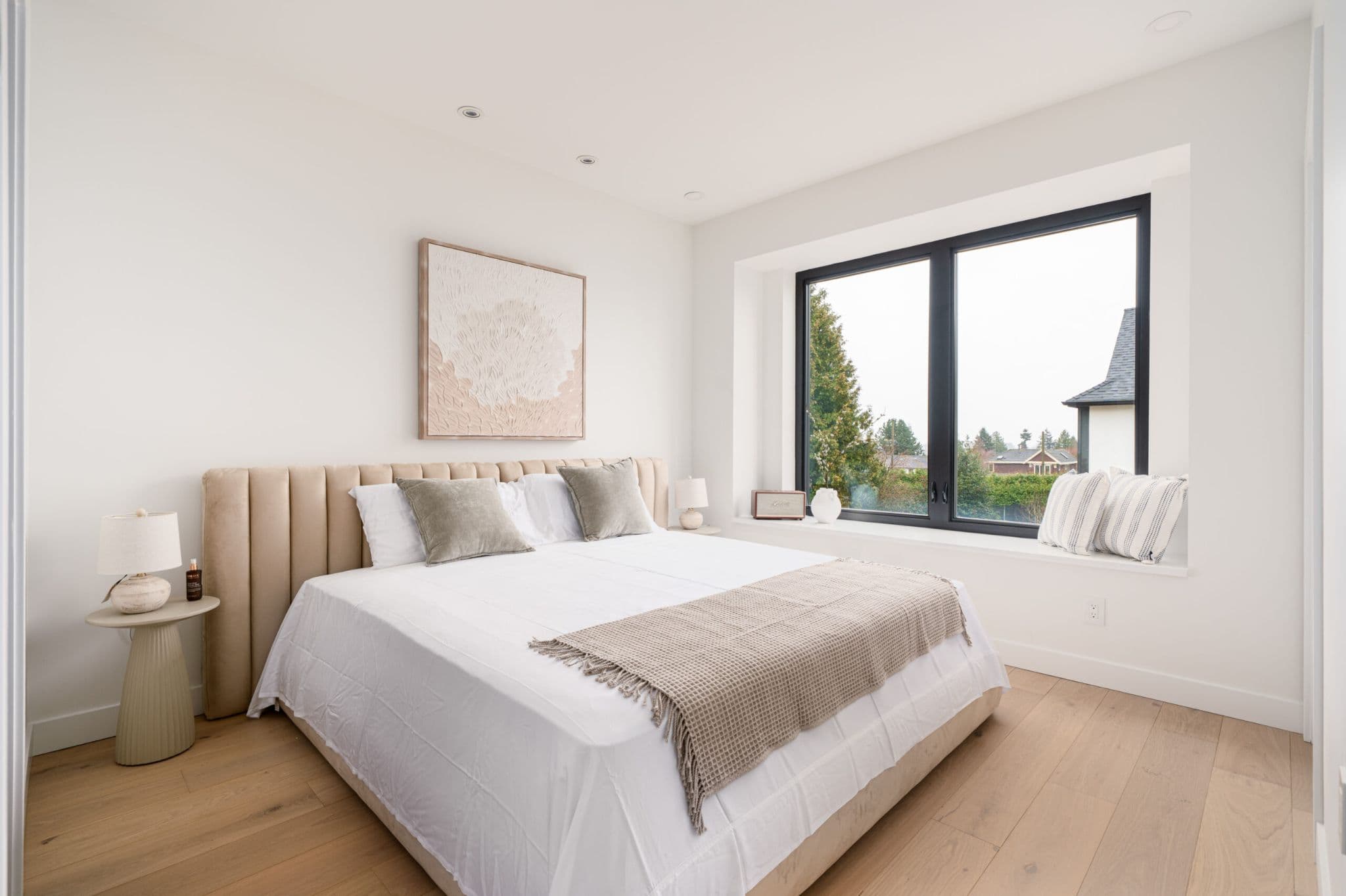 August Laurel + W64 – A modern bedroom feature a king-sized bed with a beige channeled headboard, neutral throw pillows, and light wood flooring. A large black-framed window offers a deep window sill setup with accent pillows and views of green treetops and suburban rooftops under overcast daylight.