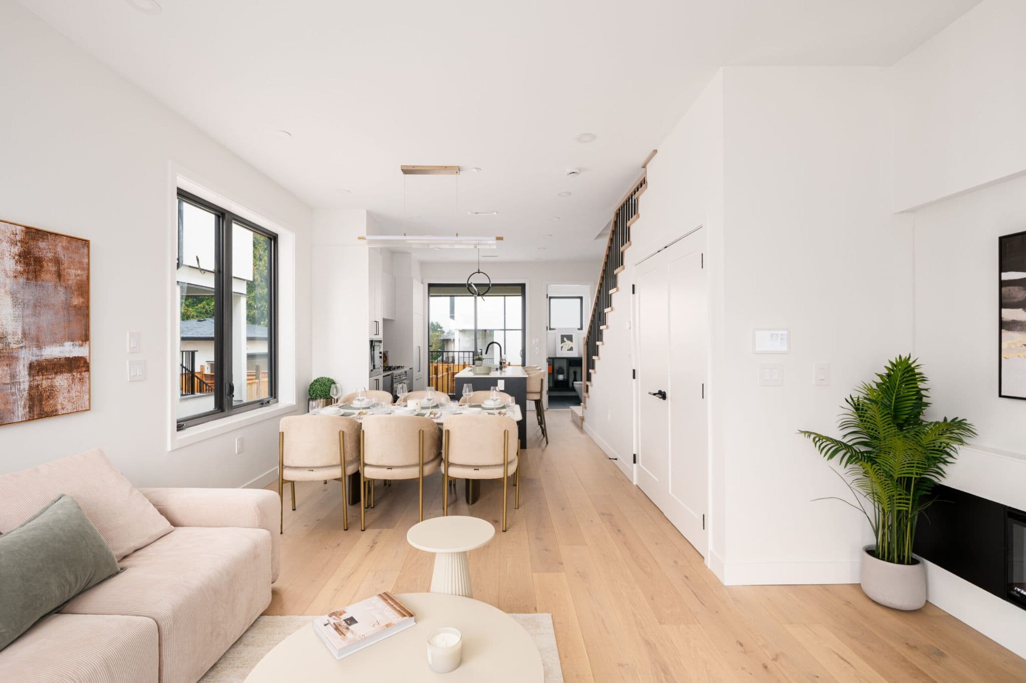 August Laurel + W64 – A modern open-concept main floor features light hardwood flooring, crisp white walls, and a living space flowing into a dining set with beige upholstered chairs and an adjacent kitchen island. Sunlight streams through large black-framed windows, highlighting clean architectural lines and an open staircase leading upstairs.