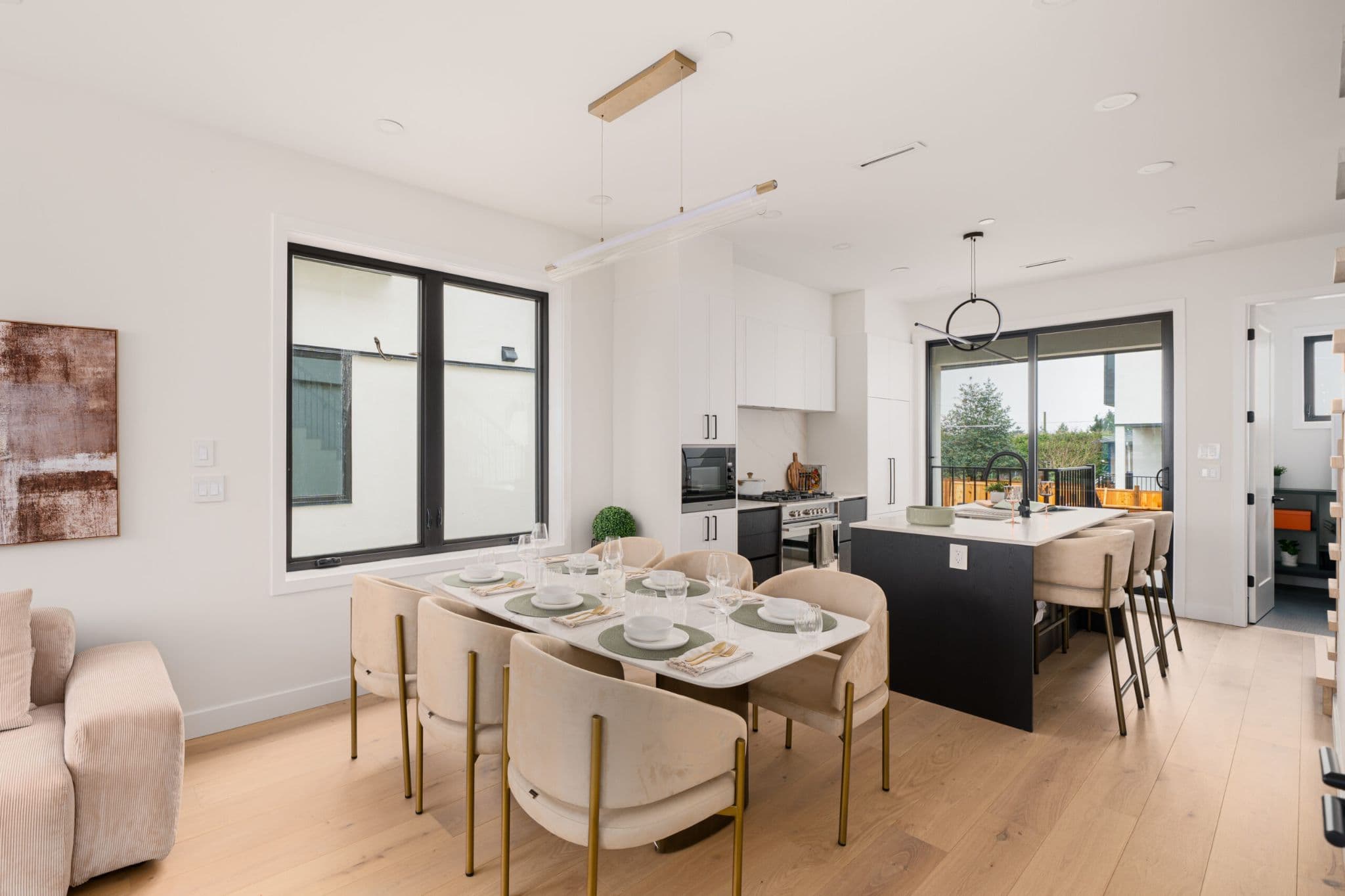 August Laurel + W64 – A bright, open-concept dining and kitchen area featuring light wood flooring, a white marble dining table surrounded by beige velvet armchairs, and a modern black island with barstool seating. Modern linear lighting hangs above the dining table, while large black-framed windows and sliding glass doors bring in natural daylight.