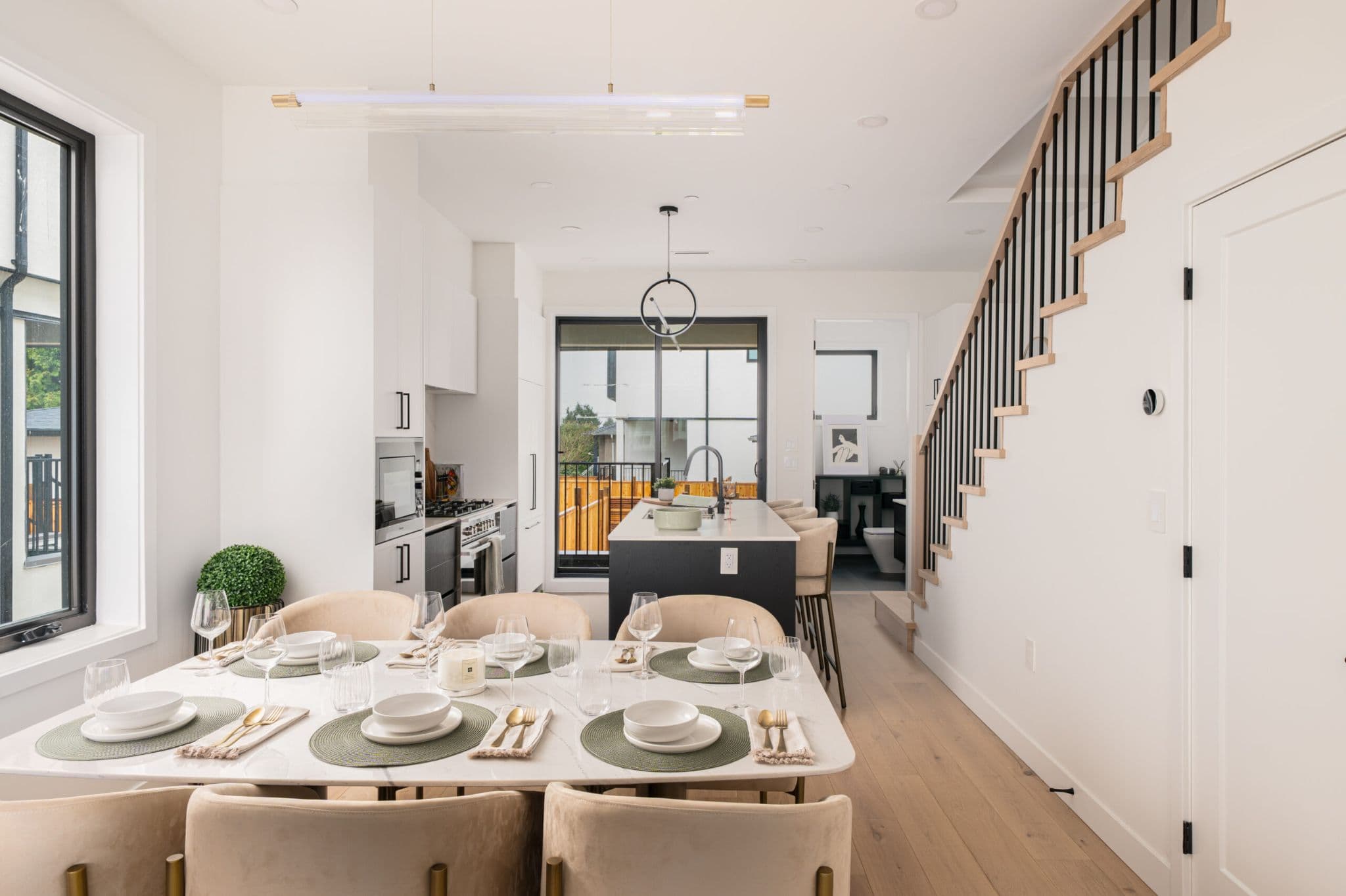 August Laurel + W64 – A modern open-concept main floor featuring a fully set marble dining table in the foreground, leading into a sleek kitchen with a dark central island and white cabinetry. Light oak wood flooring runs throughout the space, framed by bright white walls and an open staircase with black metal spindles on the right.