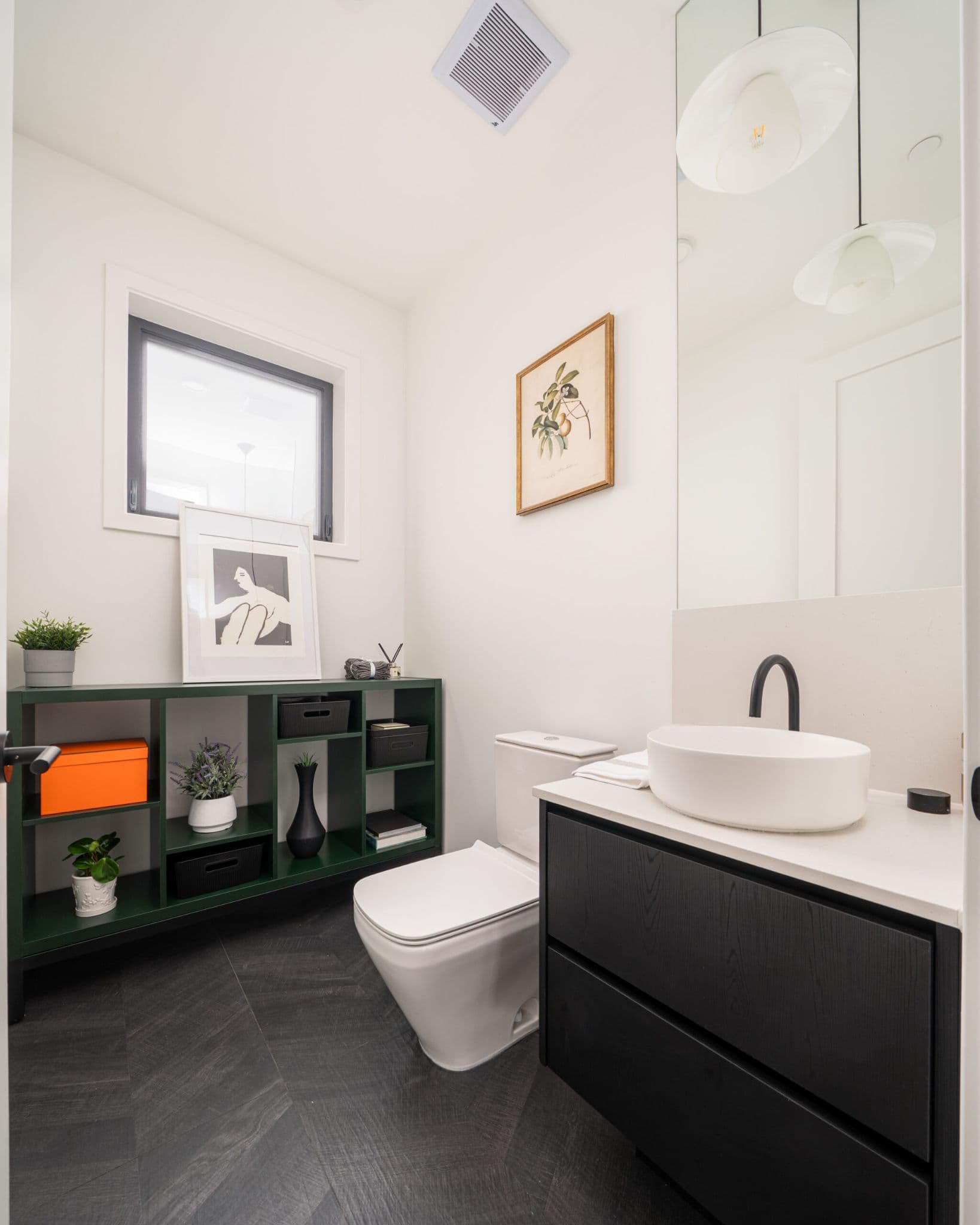 August Laurel + W64 – A modern powder room features a white oval vessel sink on a dark wood vanity with matte black fixtures, a white toilet, and dark herringbone tiled flooring. A forest green open shelving unit sits against the far wall, decorated with potted plants, storage boxes, and framed artwork.