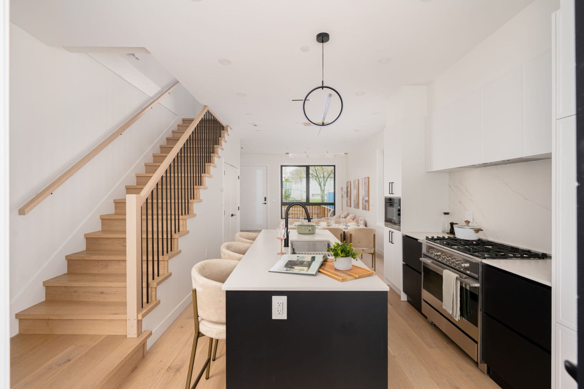 August Laurel + W64 – A modern open-concept interior featuring a bright kitchen with a black island, white quartz countertops, stainless steel gas range, and a light-wood staircase with black metal spindles leading upstairs. Bright natural light fills the space, showcasing light hardwood flooring and a dining area in the background.