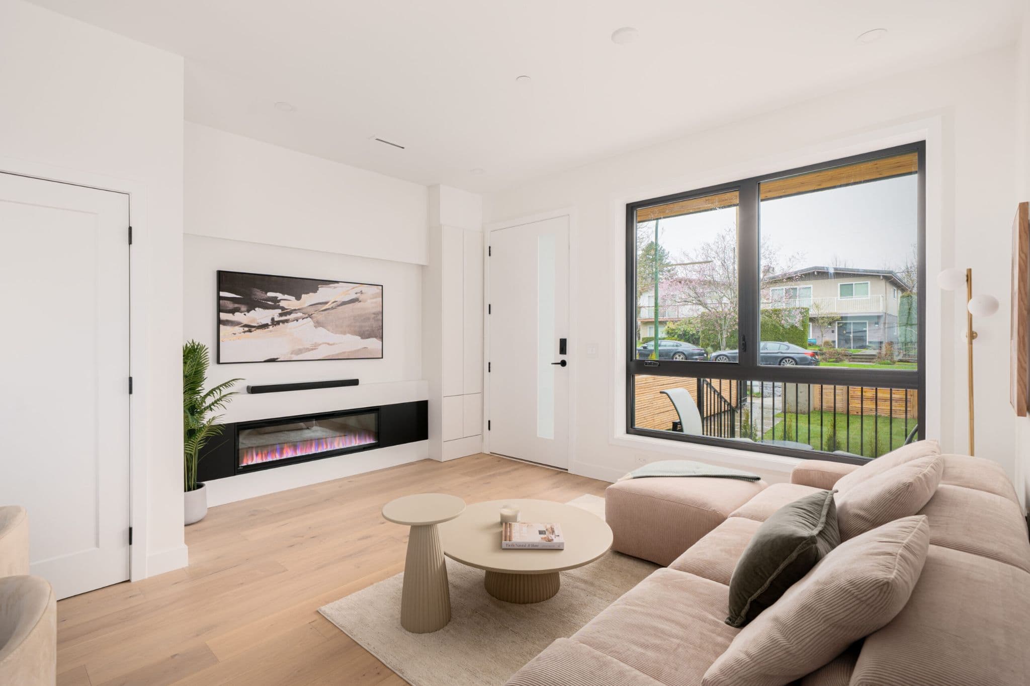 August Laurel + W64 – A modern living room features light hardwood floors, crisp white walls, and a recessed linear electric fireplace beneath a framed artwork and soundbar. A plush beige sectional sofa sits opposite a black-framed window looking out onto a residential street, complemented by round sculptural coffee tables and a potted houseplant.