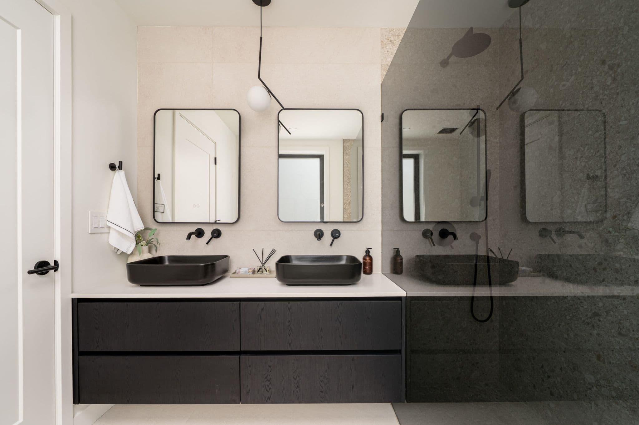 August Laurel + W64 – A modern bathroom featuring a floating dark wood vanity with dual black vessel sinks, wall-mounted matte black faucets, and two rounded rectangular mirrors. Adjacent to the vanity is a glass-enclosed shower, creating a sleek, contemporary aesthetic with neutral wall tiles and minimalist pendant lighting.