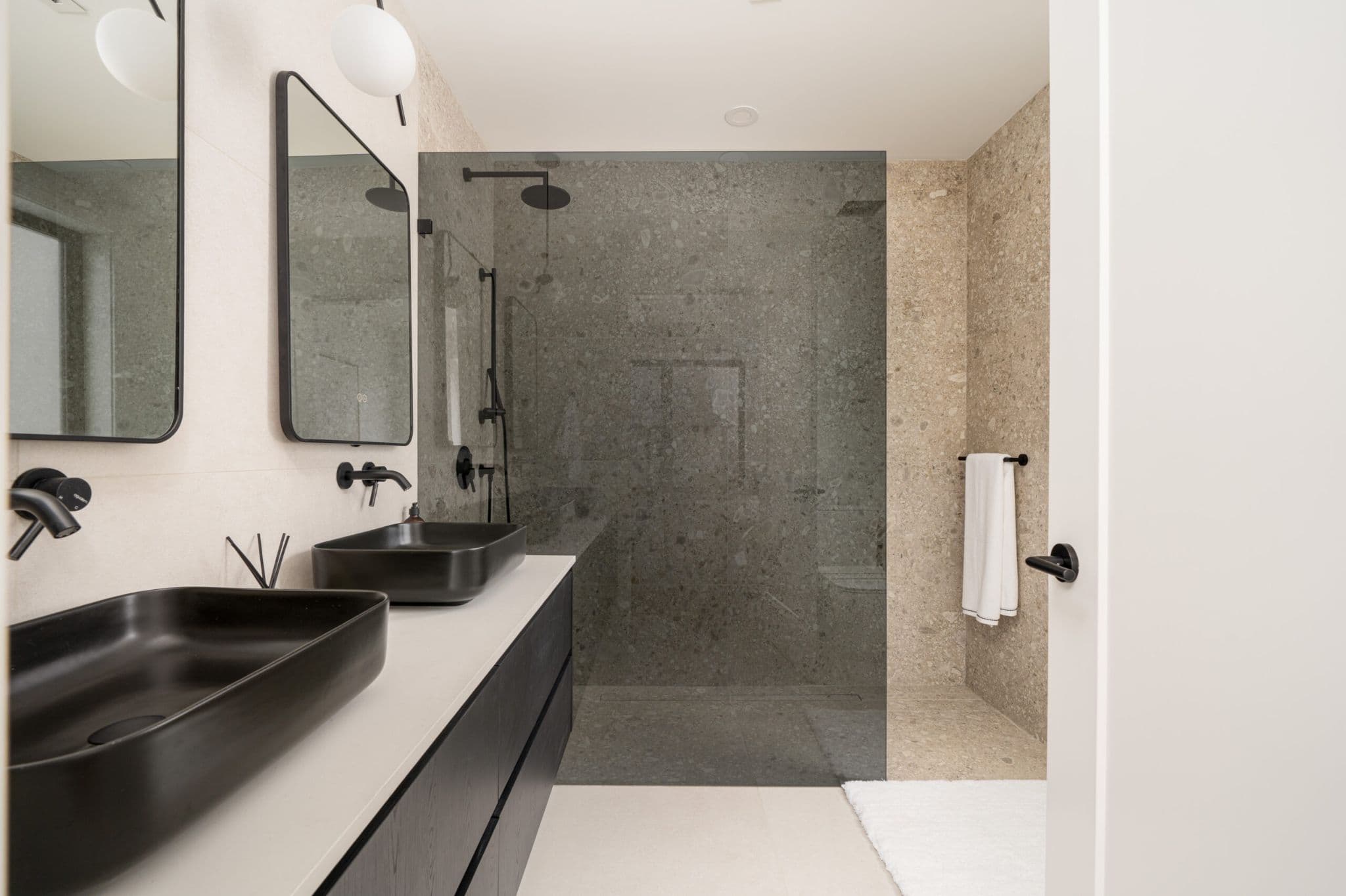 August Laurel + W64 – A modern bathroom features a double vanity with matte black vessel sinks, wall-mounted matte black faucets, and dark floating cabinetry set against light tiled walls. The adjacent walk-in shower is enclosed by glass and lined with dark grey terrazzo-style tiles, accented by matte black fixtures and warm beige stone tile accents.