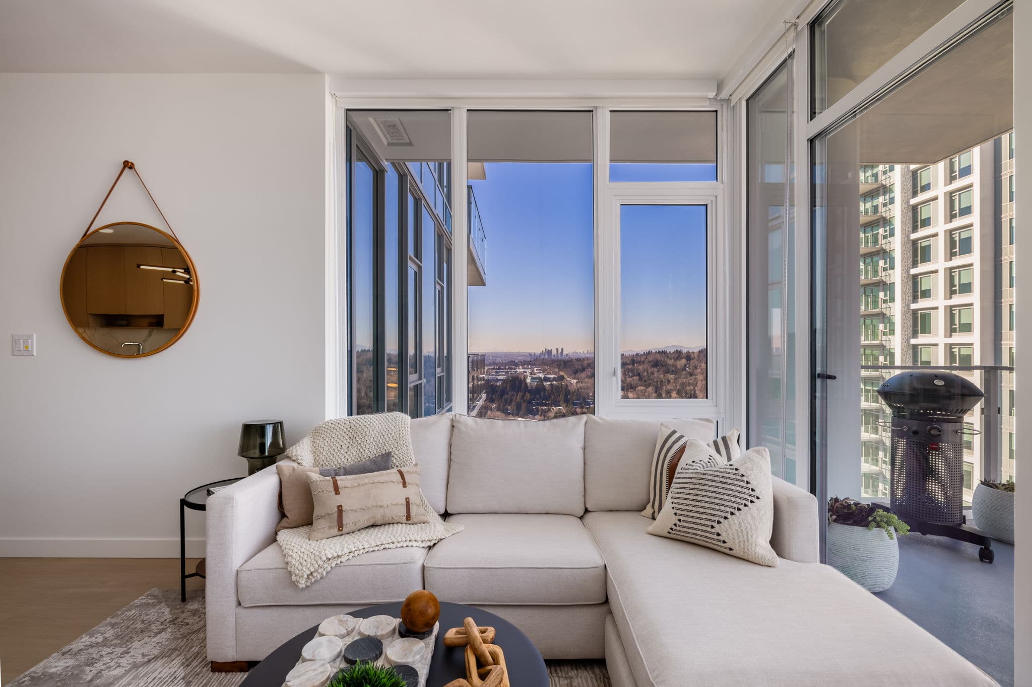 Precidia – Living room with a beige sectional facing floor-to-ceiling windows overlooking a distant city skyline, with a private balcony and grill at the side.