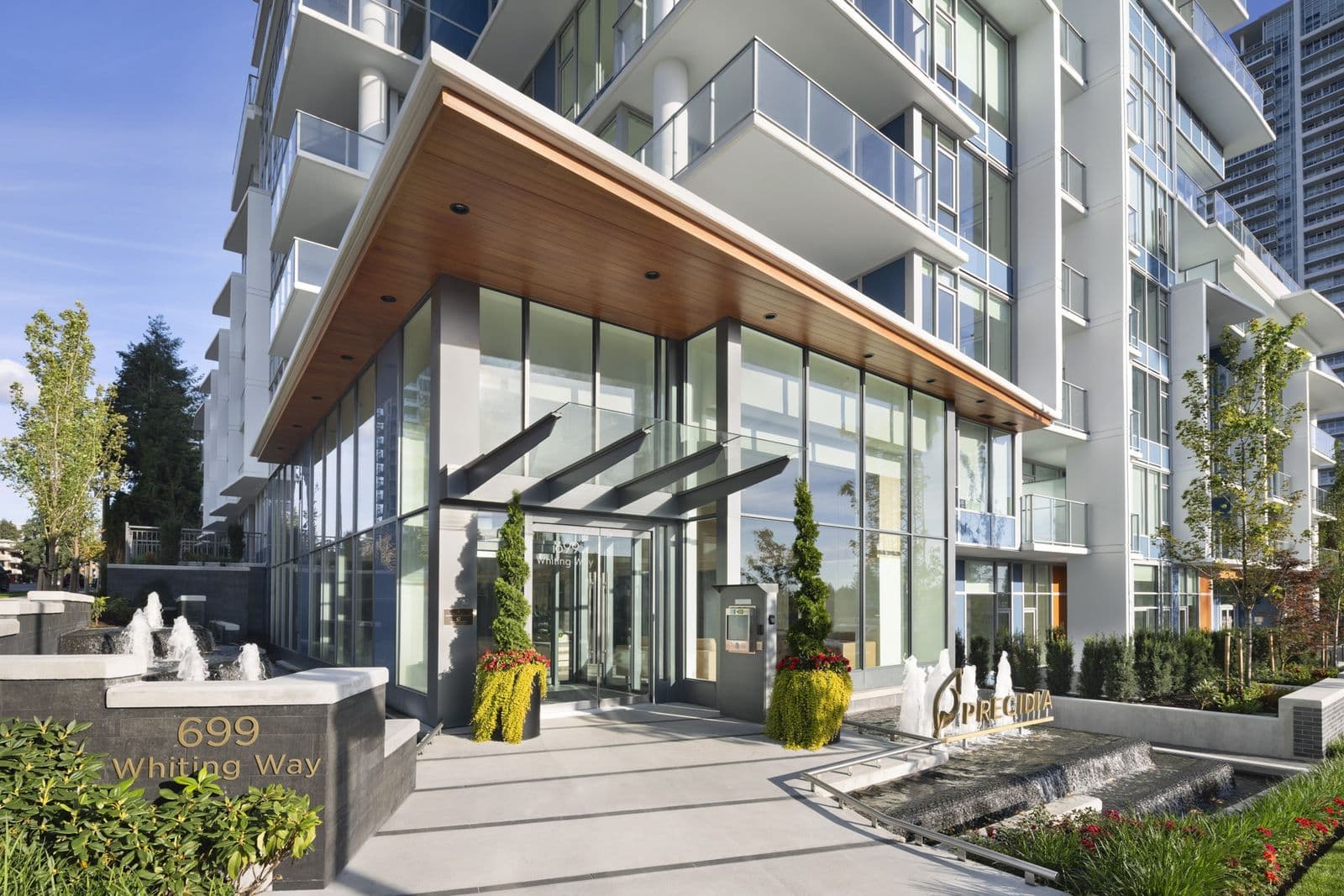 Precidia – Daytime photo of the building's entrance showing the '699 Whiting Way' address sign, glass canopy, and fountain water feature.
