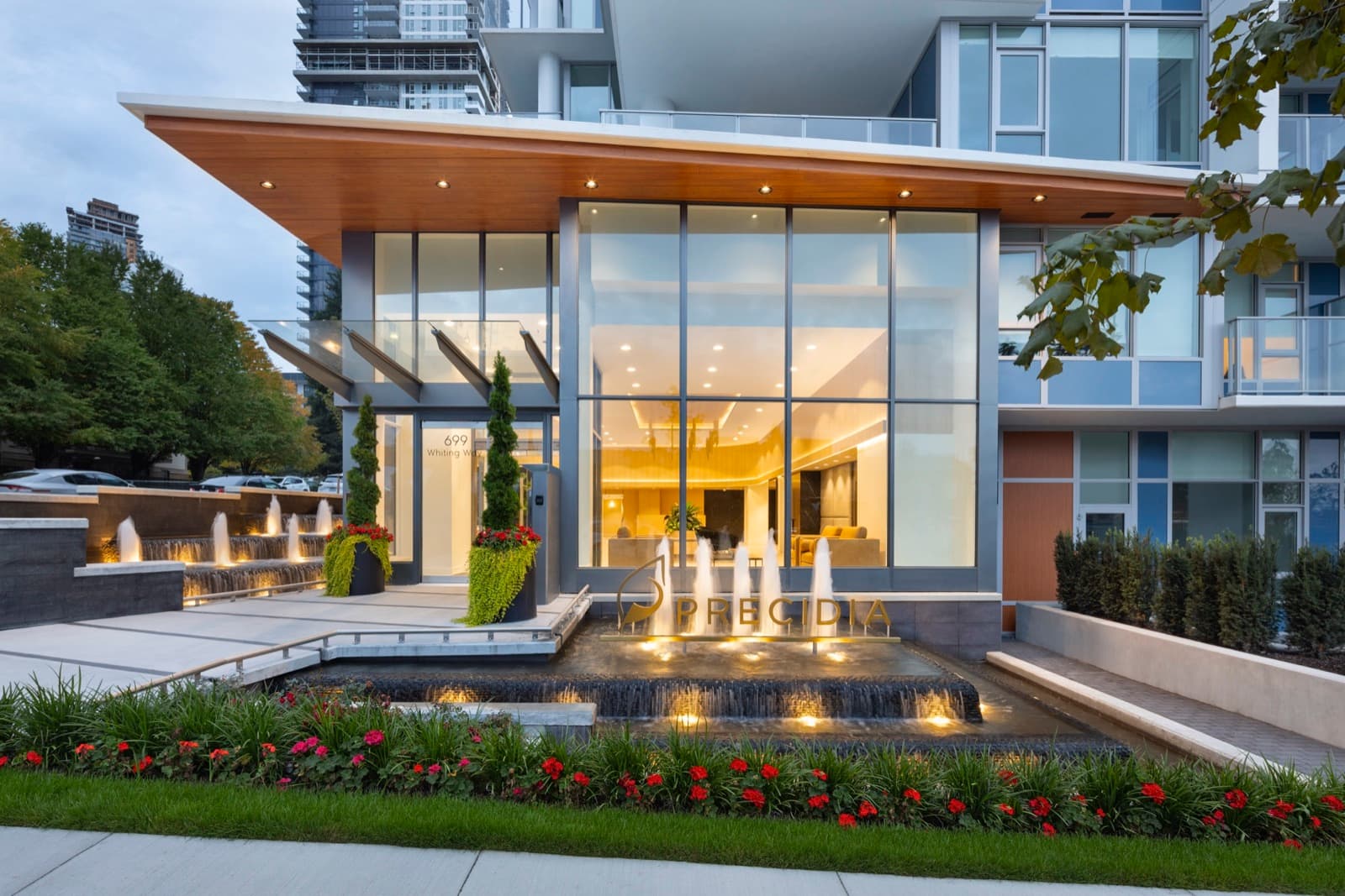 Precidia – Dusk photo of the Precidia building's glass lobby entrance with a lit water fountain feature, flower beds, and illuminated ground floor.