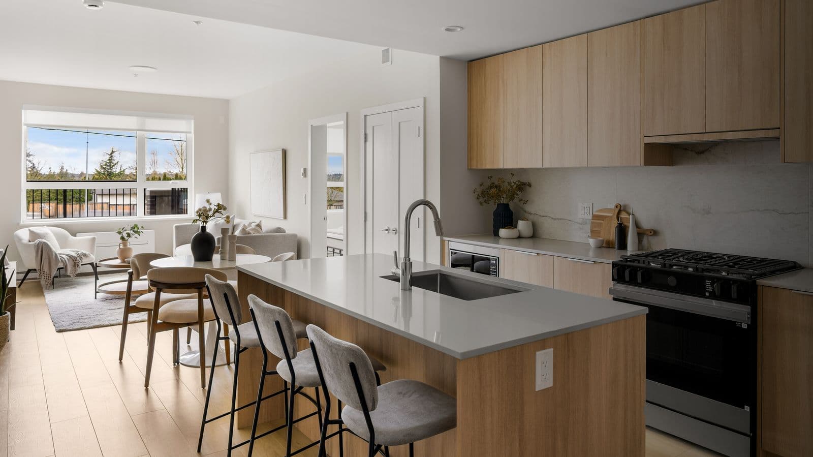 Latimer Heights – Open-concept kitchen with light wood cabinets, grey countertops, a gas range, and a view through to a living/dining area with a window overlooking trees.