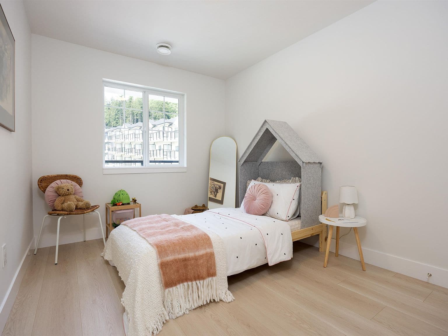 Clermont – A bright child's bedroom featuring a single bed with a playful house-shaped grey felt canopy, styled with pink and white bedding. Light wood flooring, white walls, and a window looking out onto adjacent buildings and wooded hills complete the serene, naturally lit space.