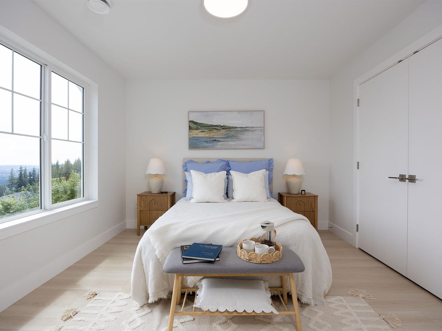 Clermont – A bright, minimalist bedroom features a neatly made bed dressed in white linens and periwinkle accent pillows, flanked by matching wooden nightstands with glowing table lamps. A bench sits at the foot of the bed with a breakfast tray, while a large window on the left frames views of lush green treetops under soft daylight.