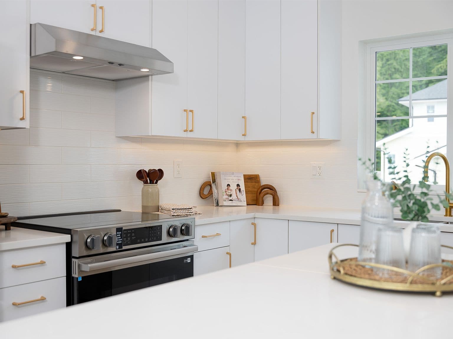 Clermont – A bright, modern kitchen featuring flat-panel white cabinetry paired with brushed gold hardware and a white subway tile backsplash with under-cabinet lighting. A stainless steel slide-in range and range hood sit beside quartz countertops, with a gold faucet visible by a window overlooking greenery.