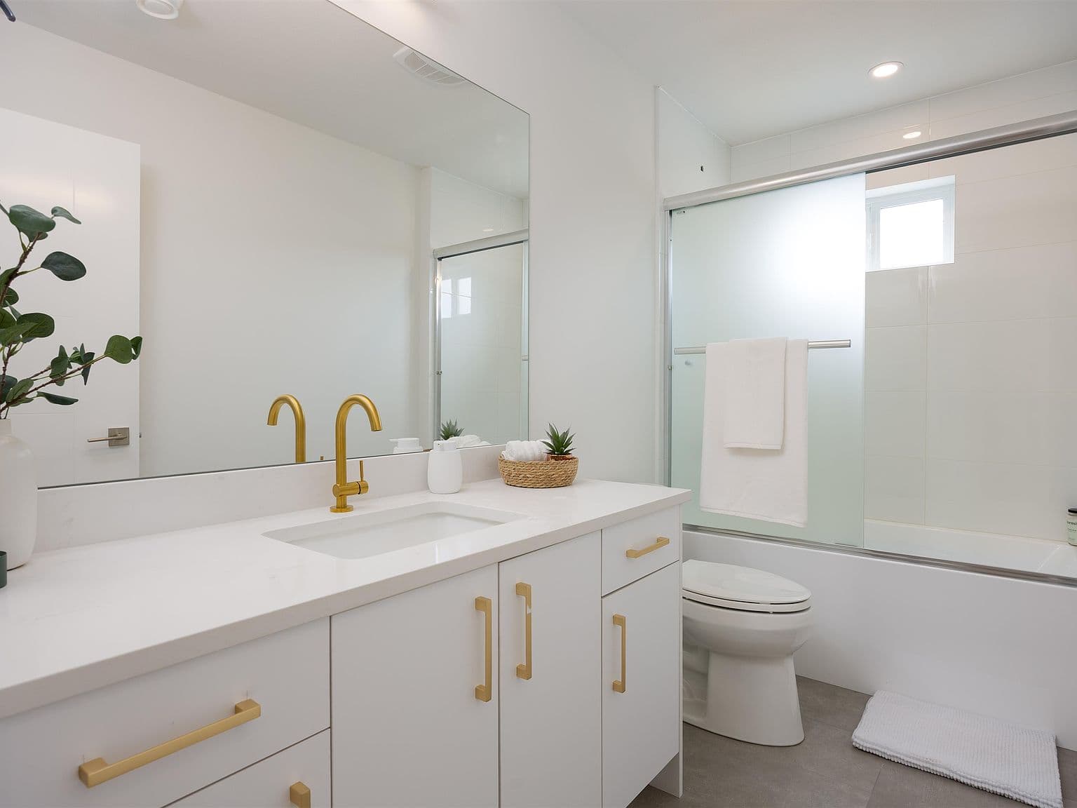 Clermont – A bright, modern bathroom featuring a white vanity with brushed gold hardware, a large mirror, and an undermount sink. Adjacent to the vanity is a toilet and a tiled bathtub and shower combination enclosed by frosted sliding glass doors.