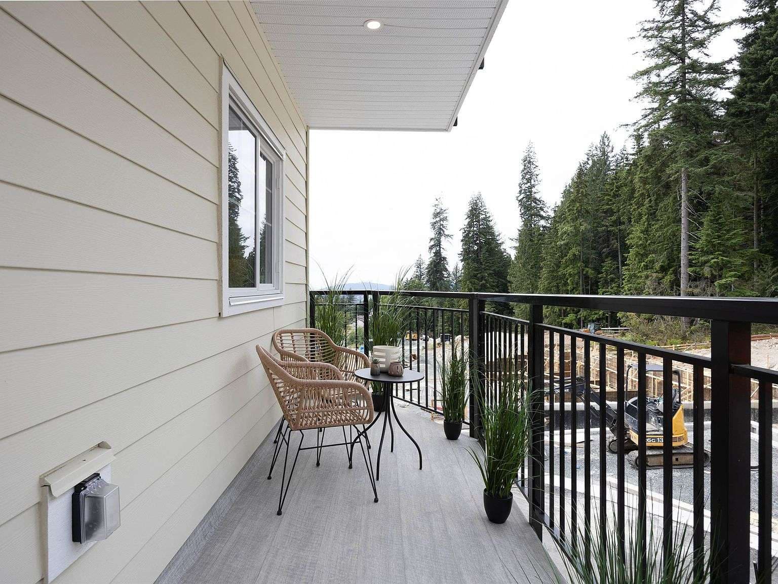 Clermont – A covered residential balcony features two woven bistro chairs, a small table, potted plants, and black metal railings looking out toward a dense evergreen forest. In the distance beyond the trees and nearby construction, soft hills and daylight skies are visible.