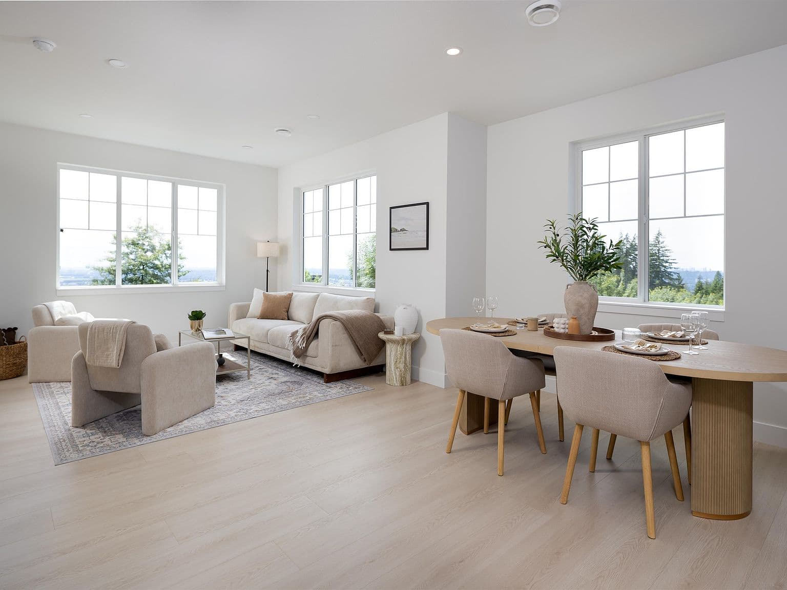 Clermont – A bright, open-concept living and dining area features light wood-look flooring, crisp white walls, and large gridded windows looking out onto lush green treetops. The space is styled with neutral beige and cream furniture, including an oval dining table with upholstered chairs and a cozy living room seating arrangement with a sofa and armchairs over an area rug.