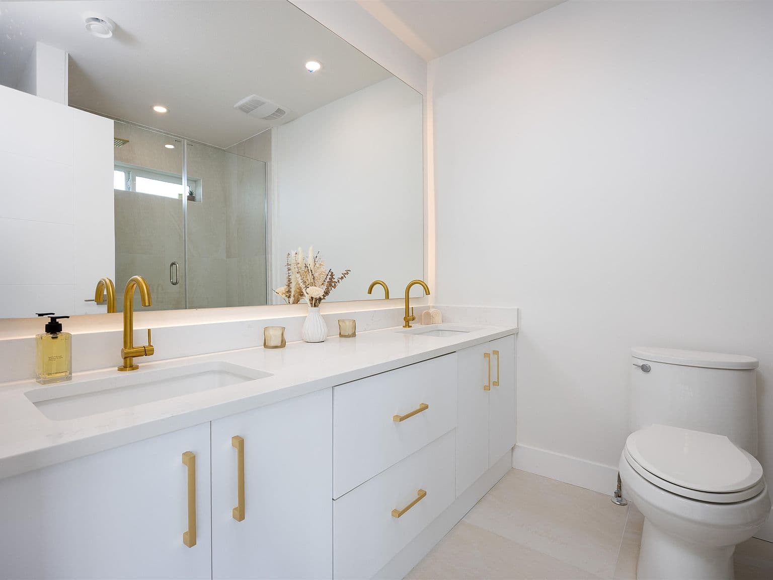 Clermont – A bright, modern bathroom featuring a white double-sink vanity with gold hardware, sleek quartz countertops, and backlighting behind a large frameless mirror. A glass-enclosed walk-in shower is visible in the mirror's reflection against clean white walls and light-toned tile flooring.