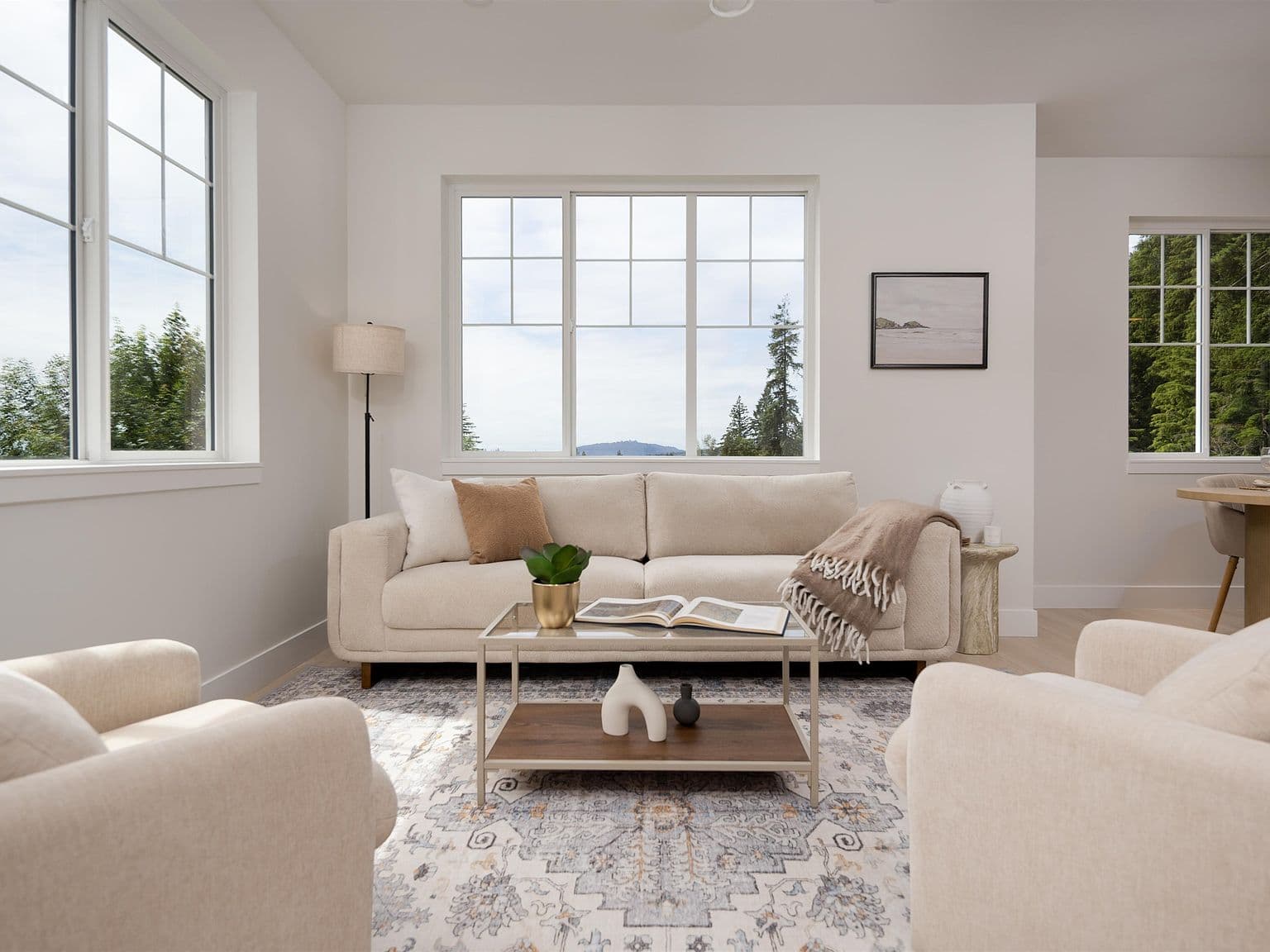 Clermont – A bright, neutrally styled living room featuring a cream-colored sofa flanked by armchairs around a glass-top coffee table on an ornate area rug. Multiple multi-pane windows fill the white-walled space with natural daylight and look out onto tree-covered hills under an overcast sky.