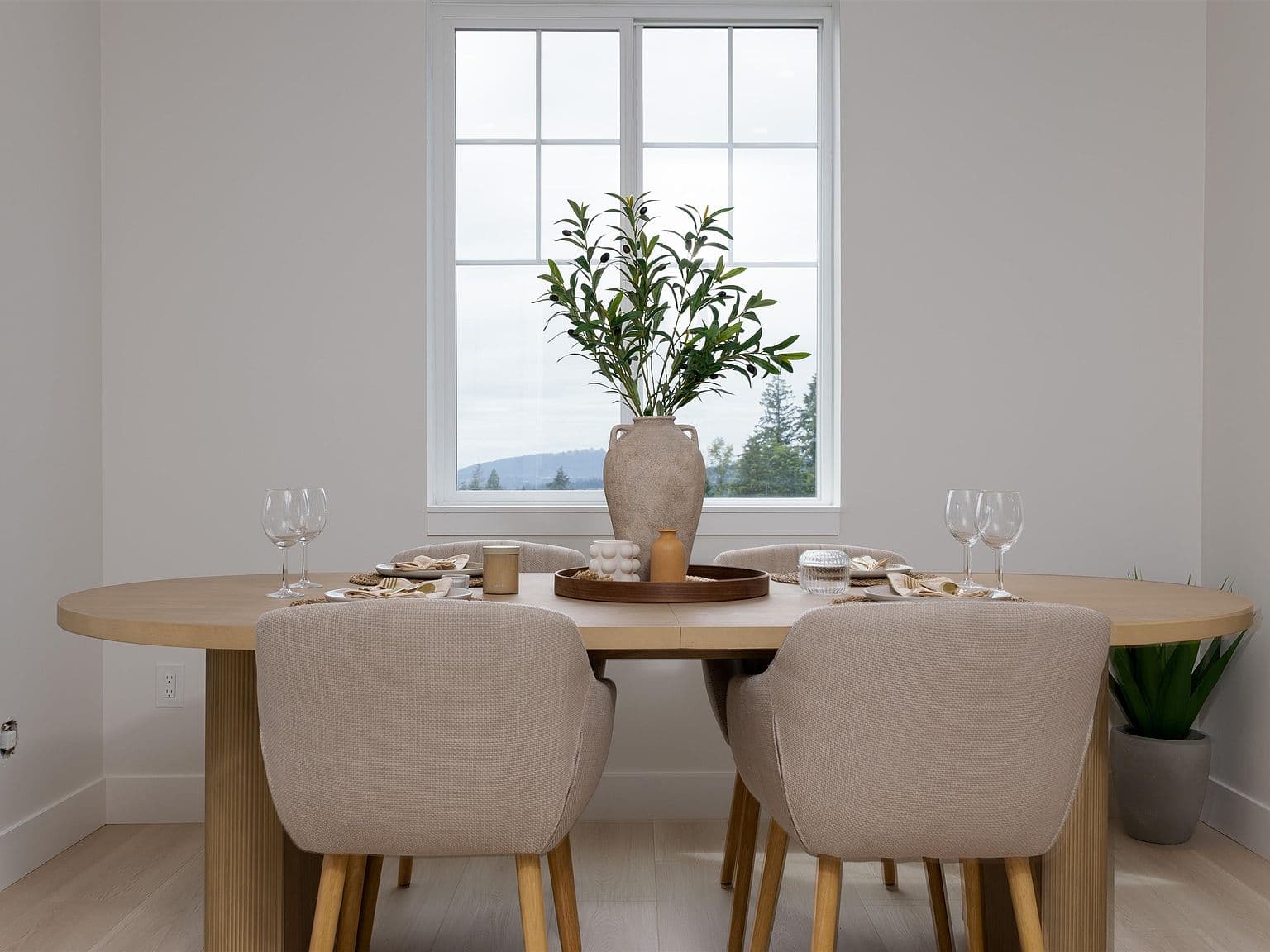 Clermont – A bright dining area features an oval light-wood dining table set with place settings, glassware, and a textured ceramic vase with greenery at its center. Upholstered beige dining chairs surround the table against white walls, with a paned window in the background looking out toward distant forested hills.