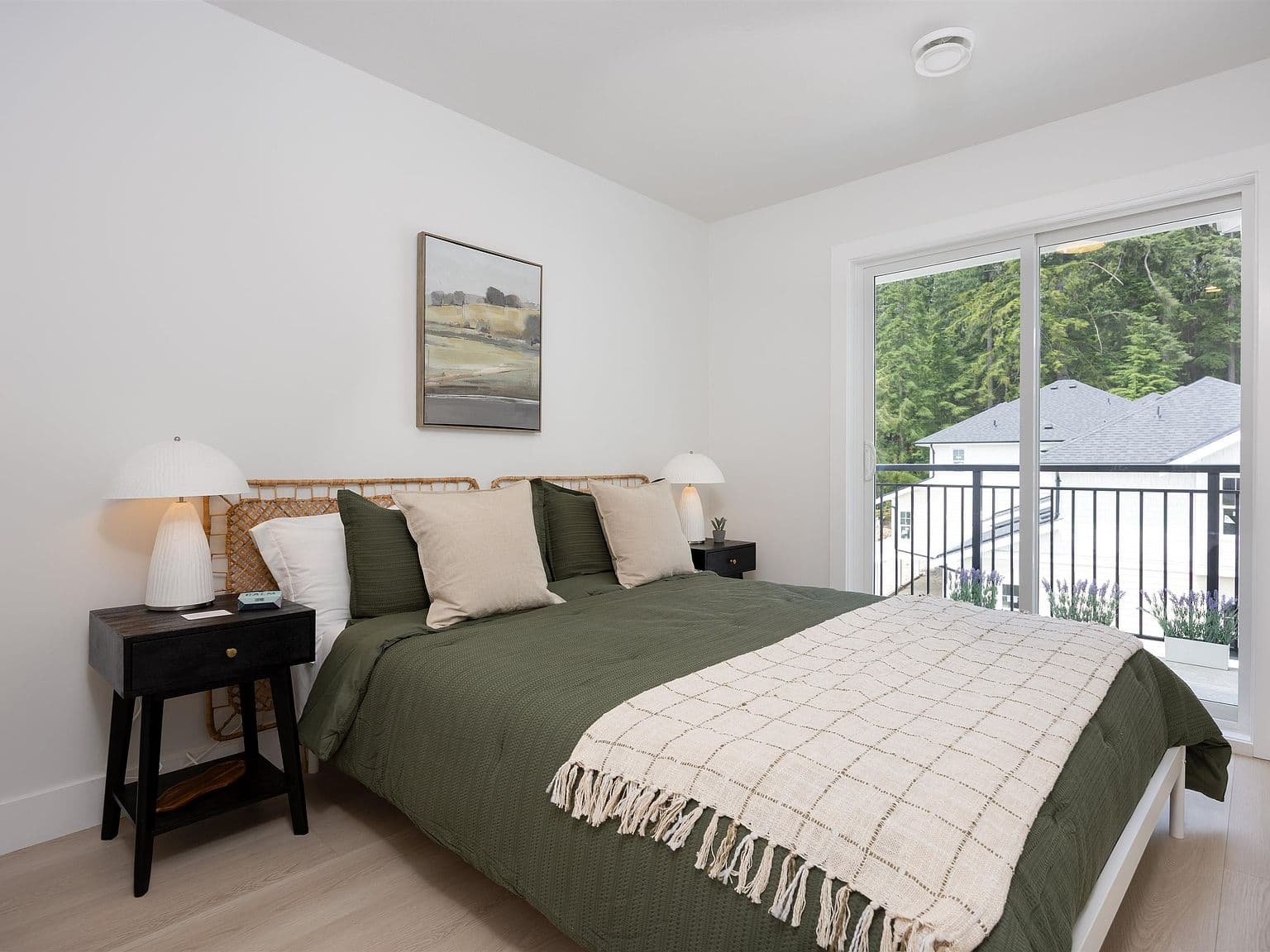 Clermont – A bright bedroom features a bed styled with olive green bedding, neutral throw pillows, a woven headboard, and matching black nightstands with lamps. Sliding glass doors lead out to a private balcony overlooking neighbouring rooftops and a lush evergreen forest.