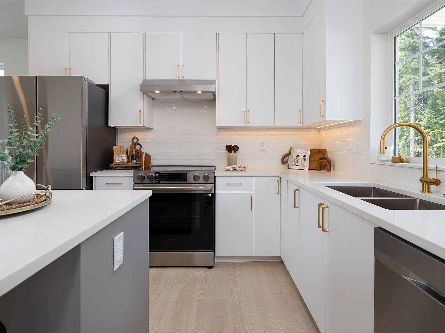 Clermont – A bright, contemporary kitchen featuring clean white cabinetry with brass hardware, white quartz countertops, and stainless steel appliances. A large window above the undermount sink with a gooseneck gold faucet lets in natural daylight and offers views of lush green trees outside.