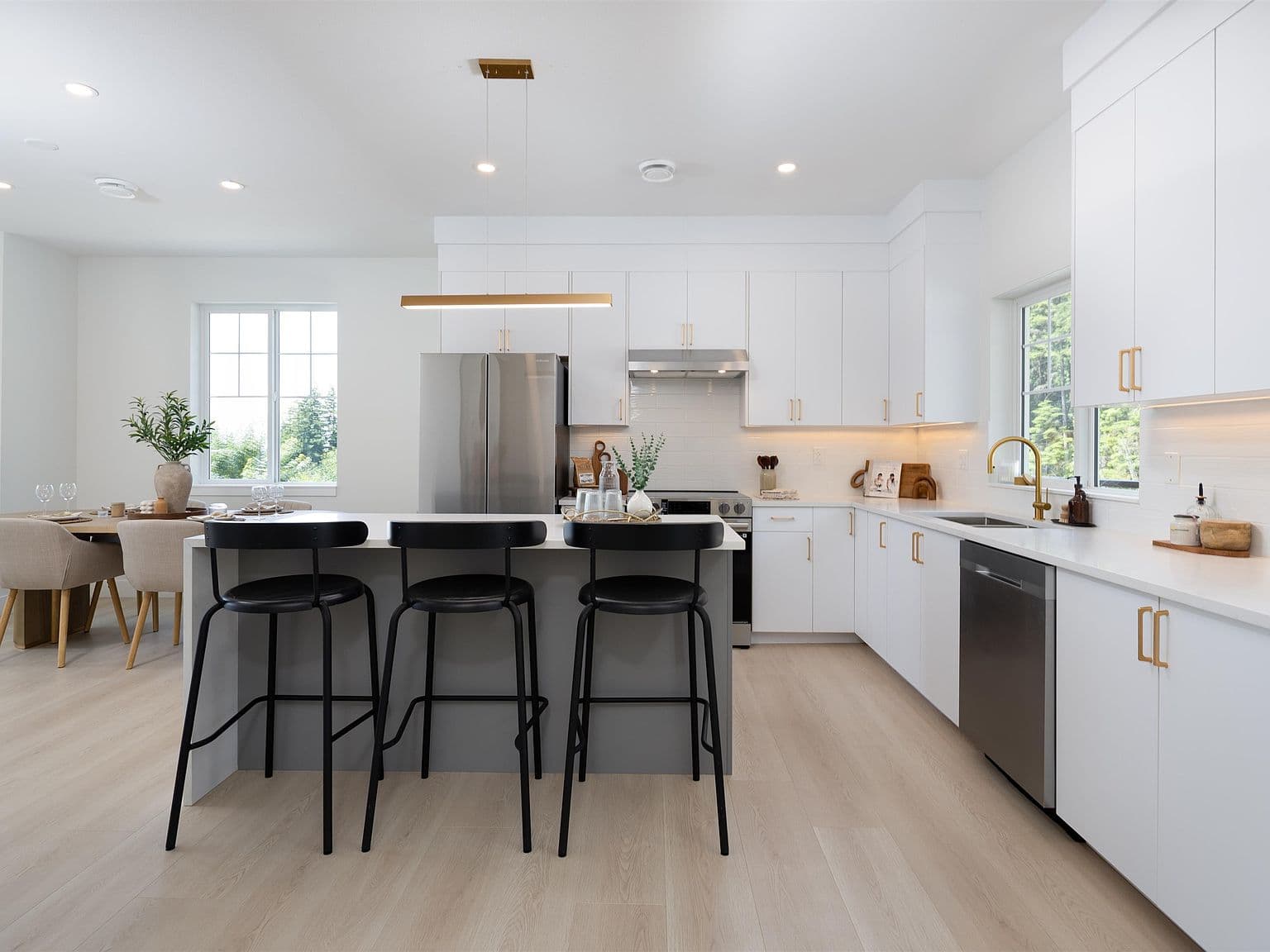 Clermont – A modern, open-concept kitchen features white flat-panel cabinetry with gold hardware, stainless steel appliances, and a central island with three black barstools. Light wood flooring runs throughout the space, which extends into an adjacent dining area beside windows letting in bright natural daylight.