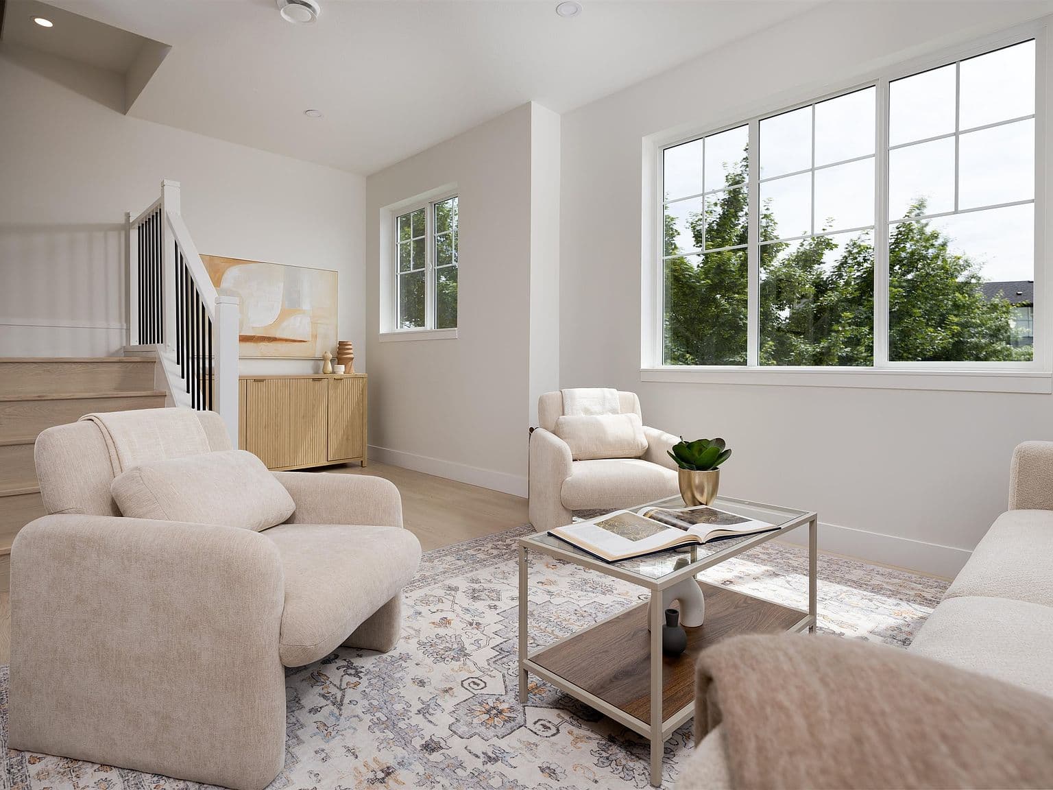 Clermont – A bright, neutrally styled living room featuring plush beige armchairs, a patterned area rug, and a glass-topped coffee table. Large multi-pane windows offer abundant natural daylight and views of lush green trees, while a wooden staircase and fluted credenza are visible in the background.