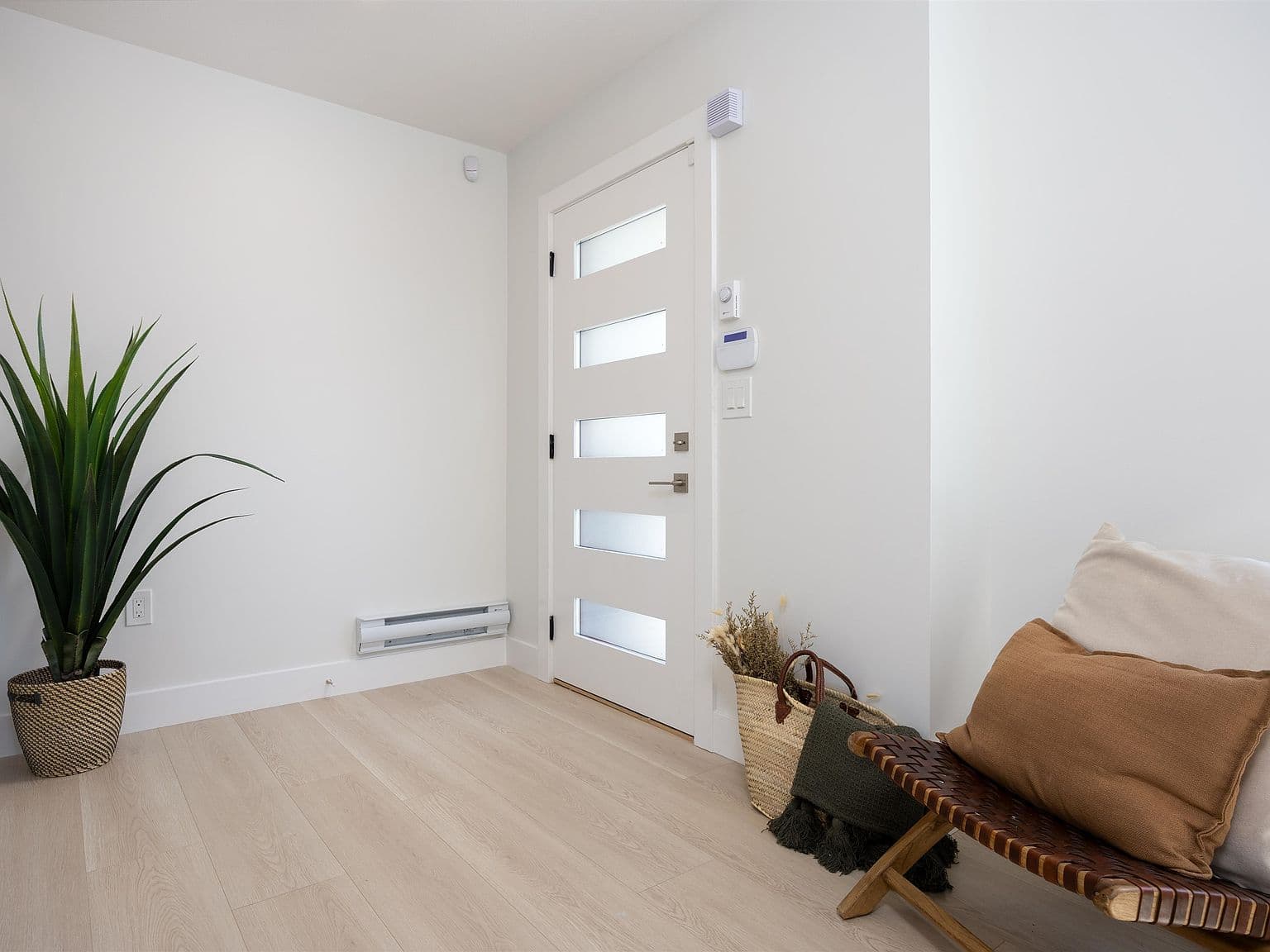 Clermont – A bright home entryway featuring light oak-toned flooring, white walls, and a modern white front door with five frosted glass panel inserts. Simple styling includes a tall potted plant on the left and a woven leather bench with accent pillows and a tote bag on the right.