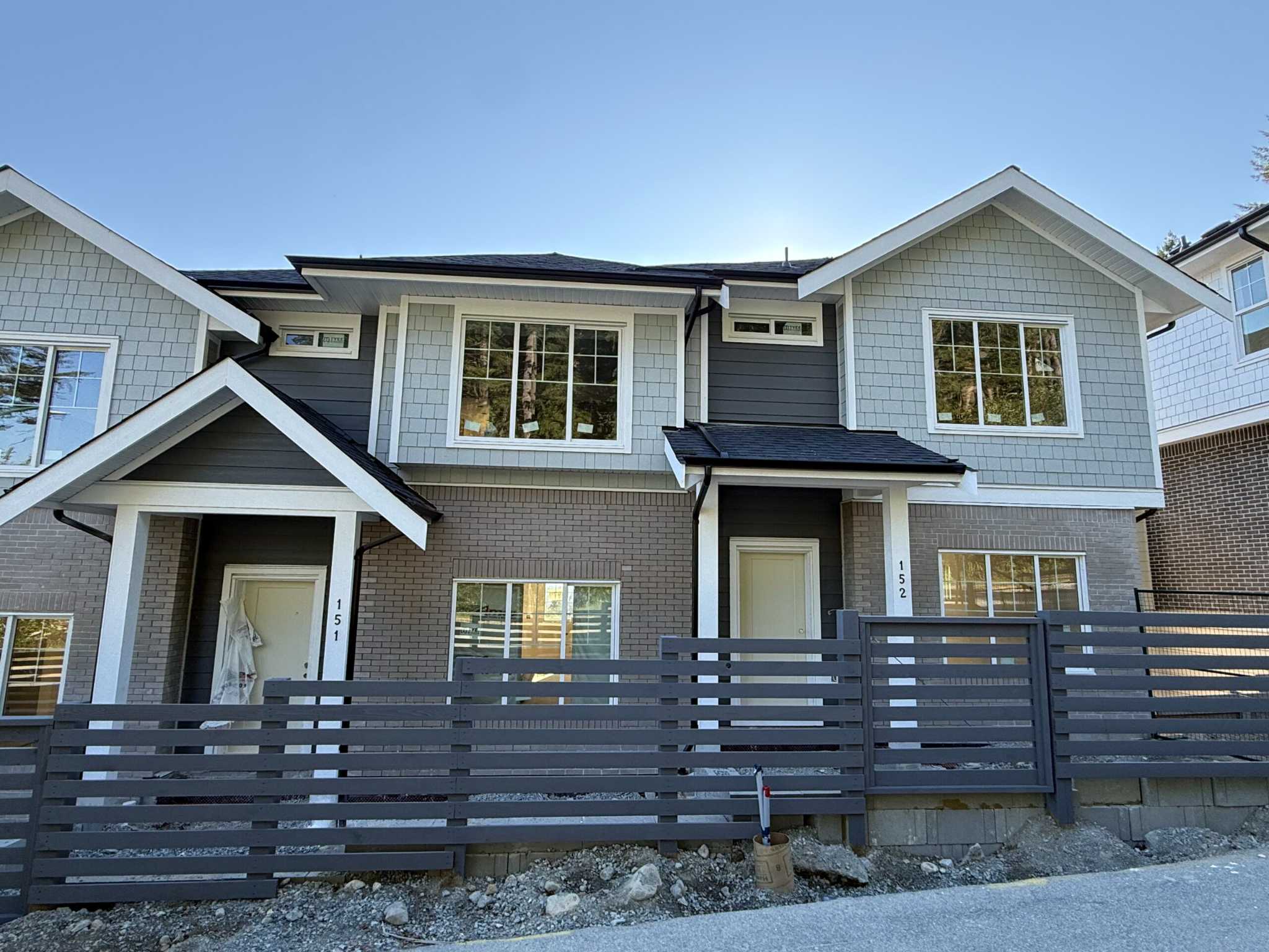 Clermont – A ground-level view of newly constructed townhomes featuring grey brick on the lower level, sage-green shake and dark siding above, and gabled rooflines under a clear blue sky. A dark horizontal slat fence lines the unpaved front yard and road in the foreground.