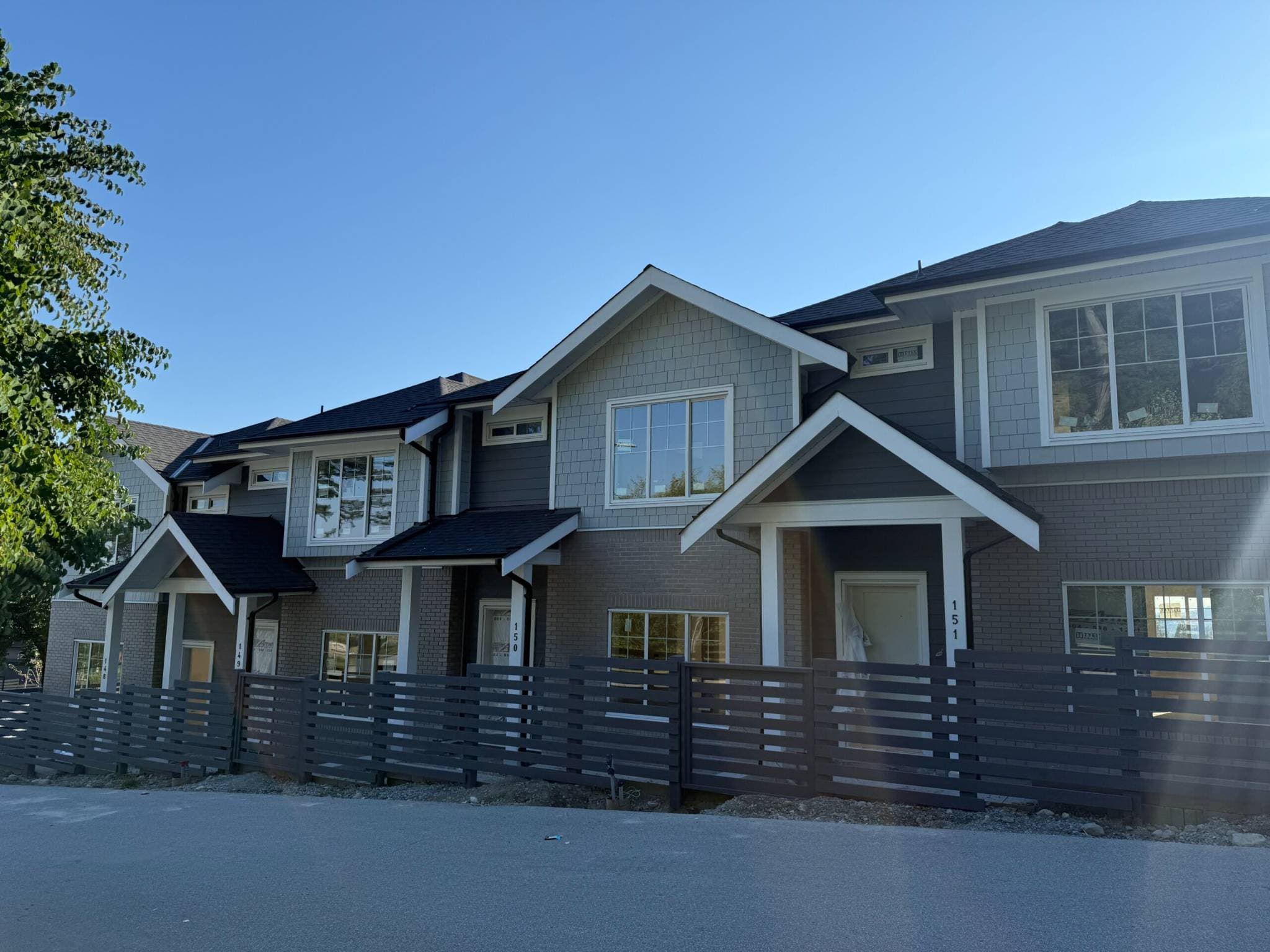 Clermont – A ground-level street view of newly constructed, multi-unit townhomes featuring grey siding, shake accents, and dark grey brick finishes. Each unit includes gabled entry porches fronted by horizontal dark-slat fencing along an unpaved edge and paved road under a clear blue sky.