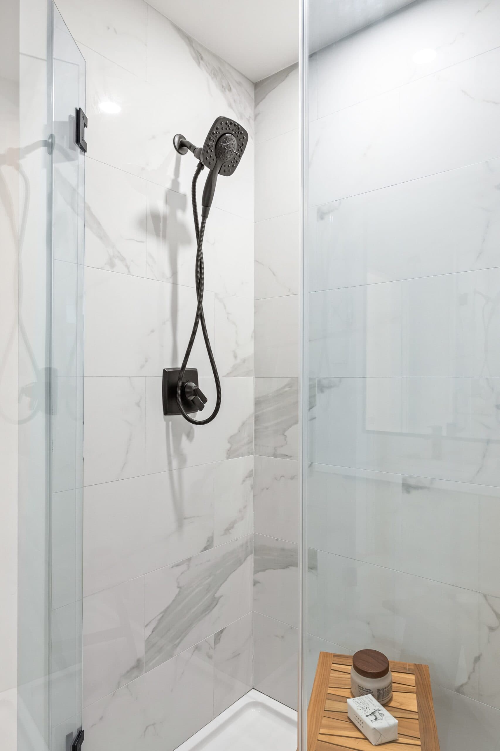 Archer at Latimer – Glass-enclosed shower with a handheld shower head and marble-look tile walls, a small wood bench with toiletries.