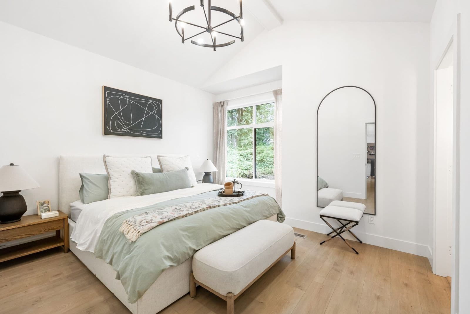 Archer at Latimer – Bedroom with an upholstered bed, sage green bedding, abstract wall art, and an arched full-length mirror by the window.