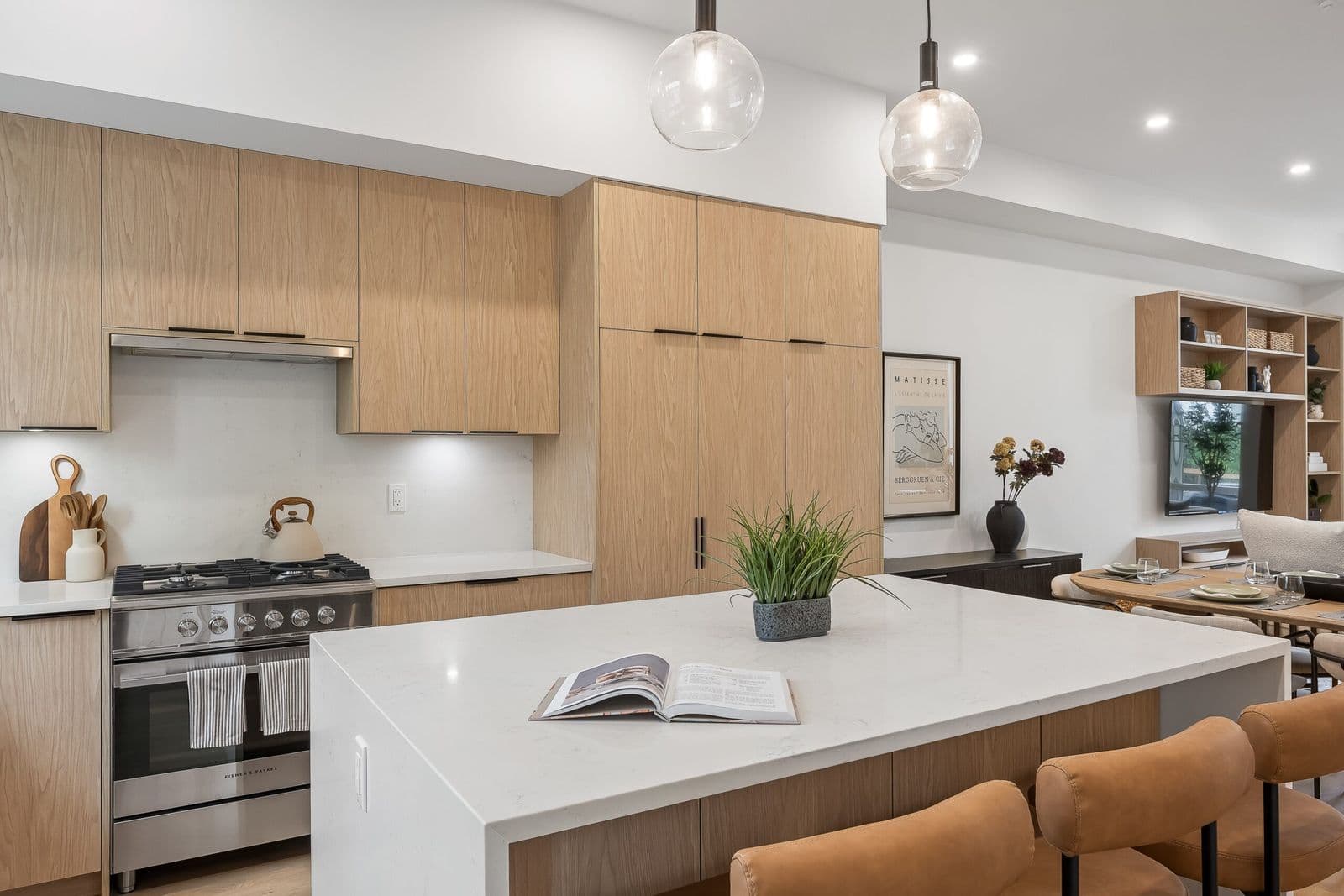 Archer at Latimer – Kitchen with light wood cabinetry, white quartz island, gas range, and glass pendant lights, opening to a living area.